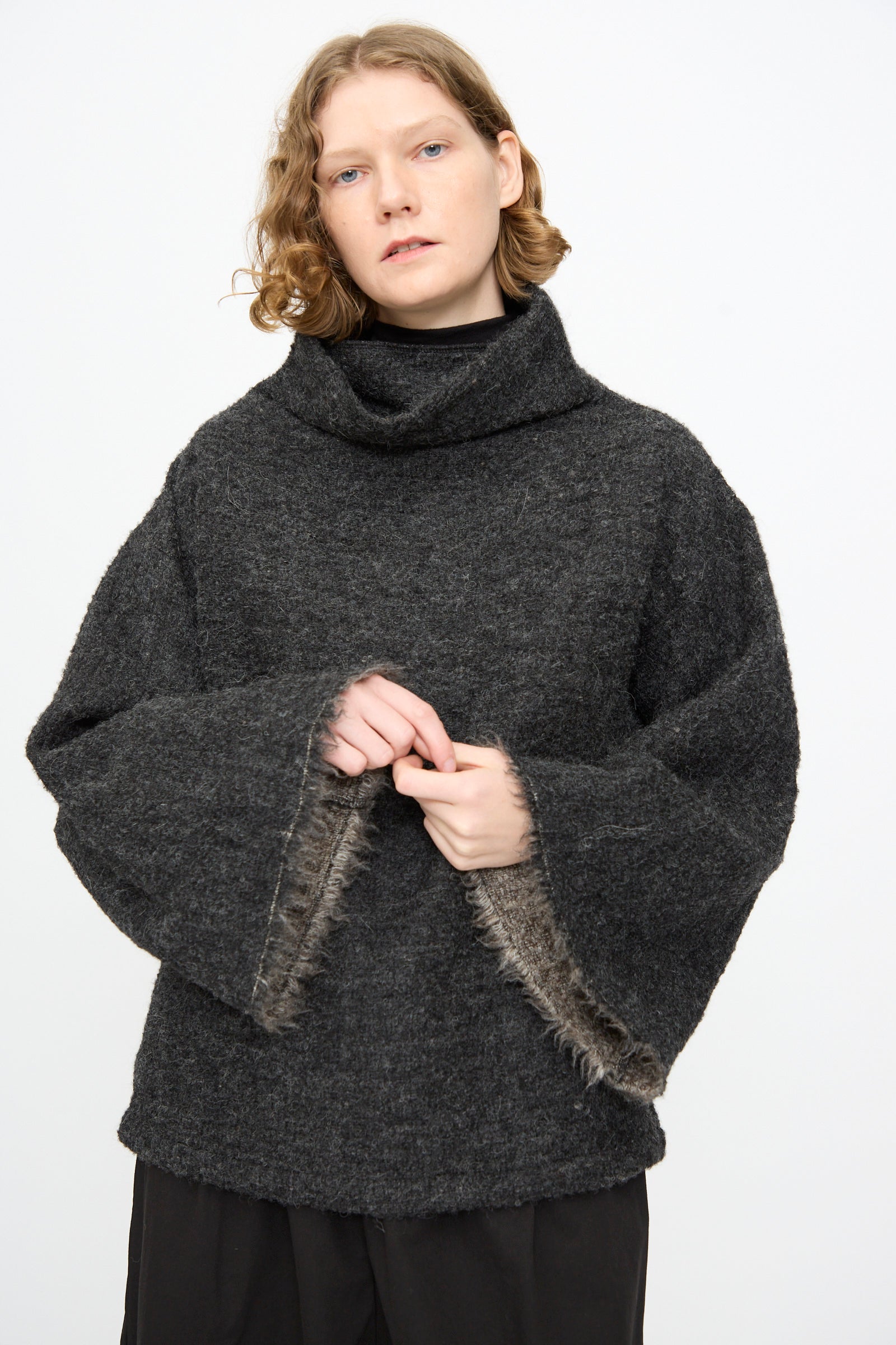 Wearing the Black Crane Alpaca Tube Top in Dark Grey, a person with short, wavy hair stands against a plain white background, layered under a loose sweater with wide sleeves and fur trim.