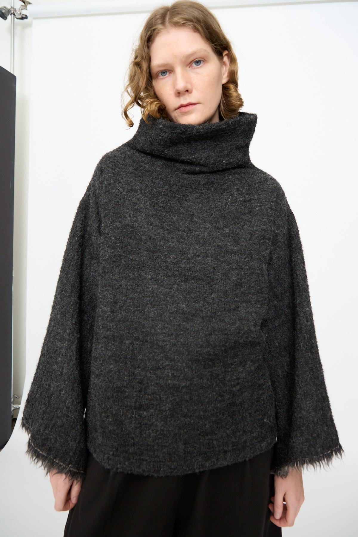 A person with wavy, shoulder-length hair is wearing the Black Crane Alpaca Tube Top in Dark Grey, featuring a loose fit and fringed hem, paired with black pants and standing against a plain white background.