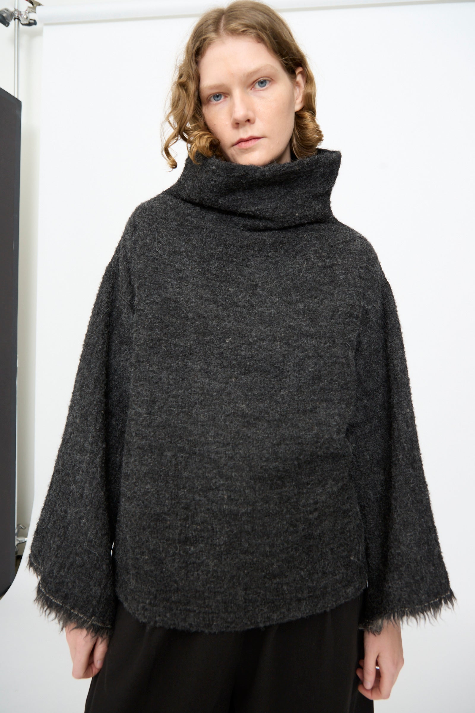 A person with wavy, shoulder-length hair is wearing the Black Crane Alpaca Tube Top in Dark Grey, featuring a loose fit and fringed hem, paired with black pants and standing against a plain white background.