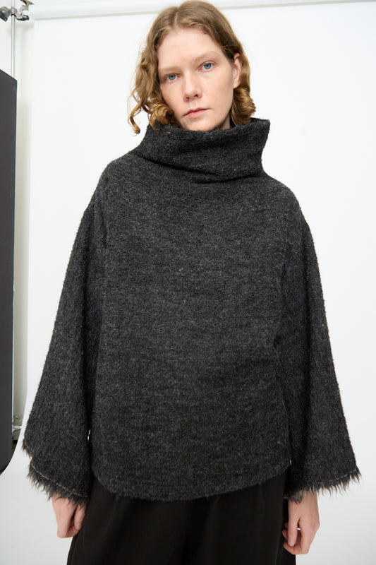 A person with wavy, shoulder-length hair is wearing the Black Crane Alpaca Tube Top in Dark Grey, featuring a loose fit and fringed hem, paired with black pants and standing against a plain white background.