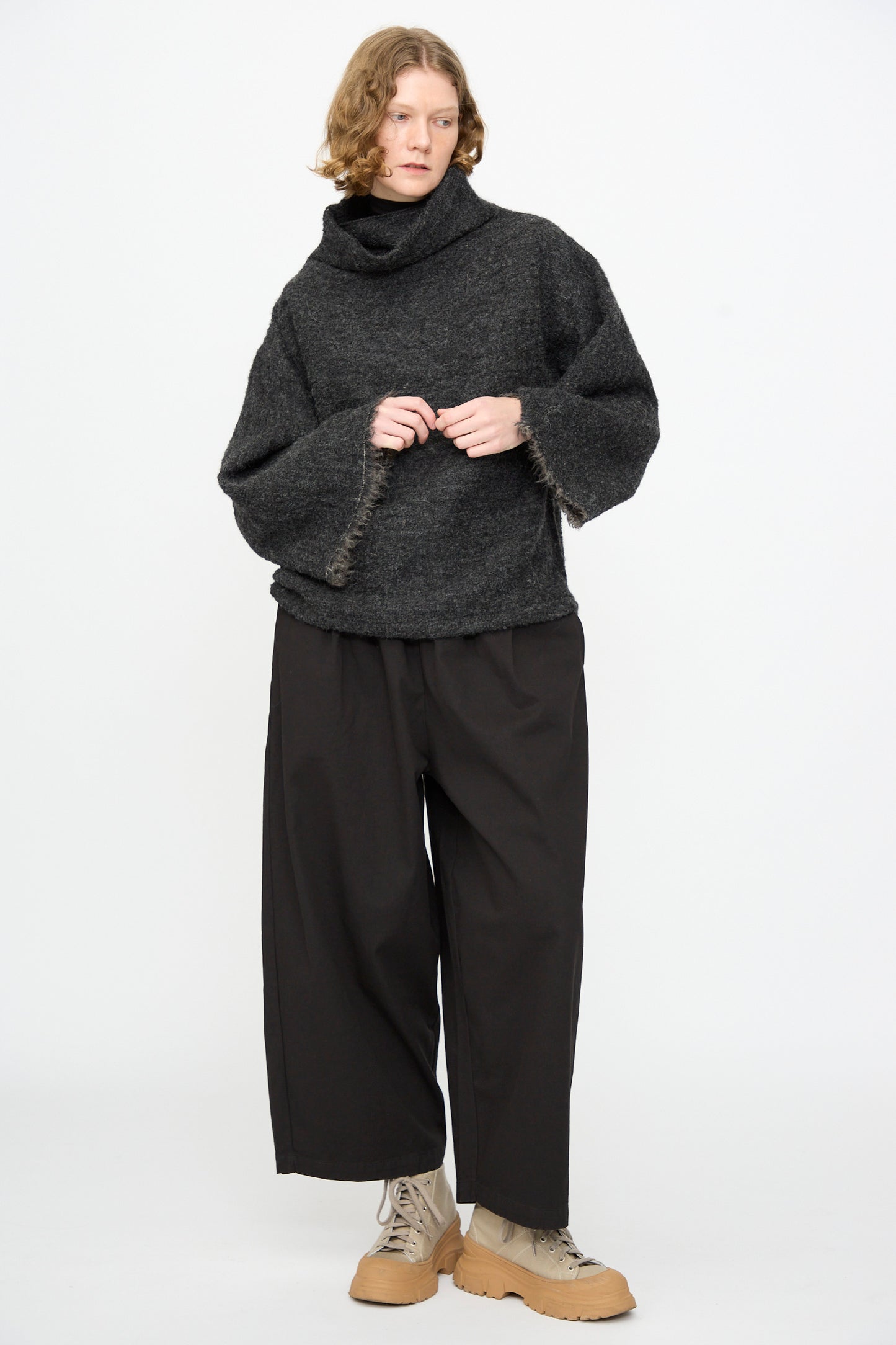 A person stands against a white background wearing the Black Crane Alpaca Tube Top in Dark Grey styled as a turtleneck sweater, paired with wide-leg black pants and light brown lace-up boots.