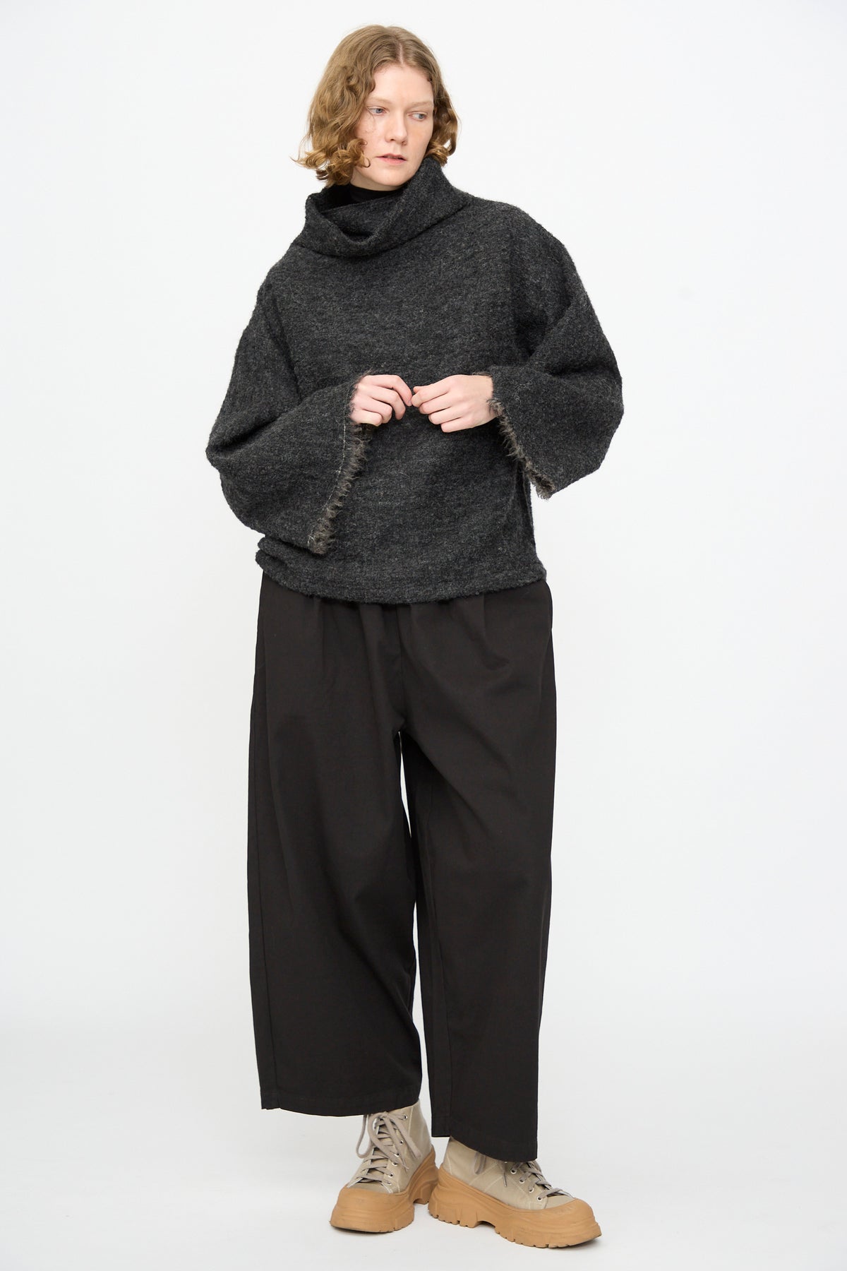 A person stands against a white background wearing the Black Crane Alpaca Tube Top in Dark Grey styled as a turtleneck sweater, paired with wide-leg black pants and light brown lace-up boots.
