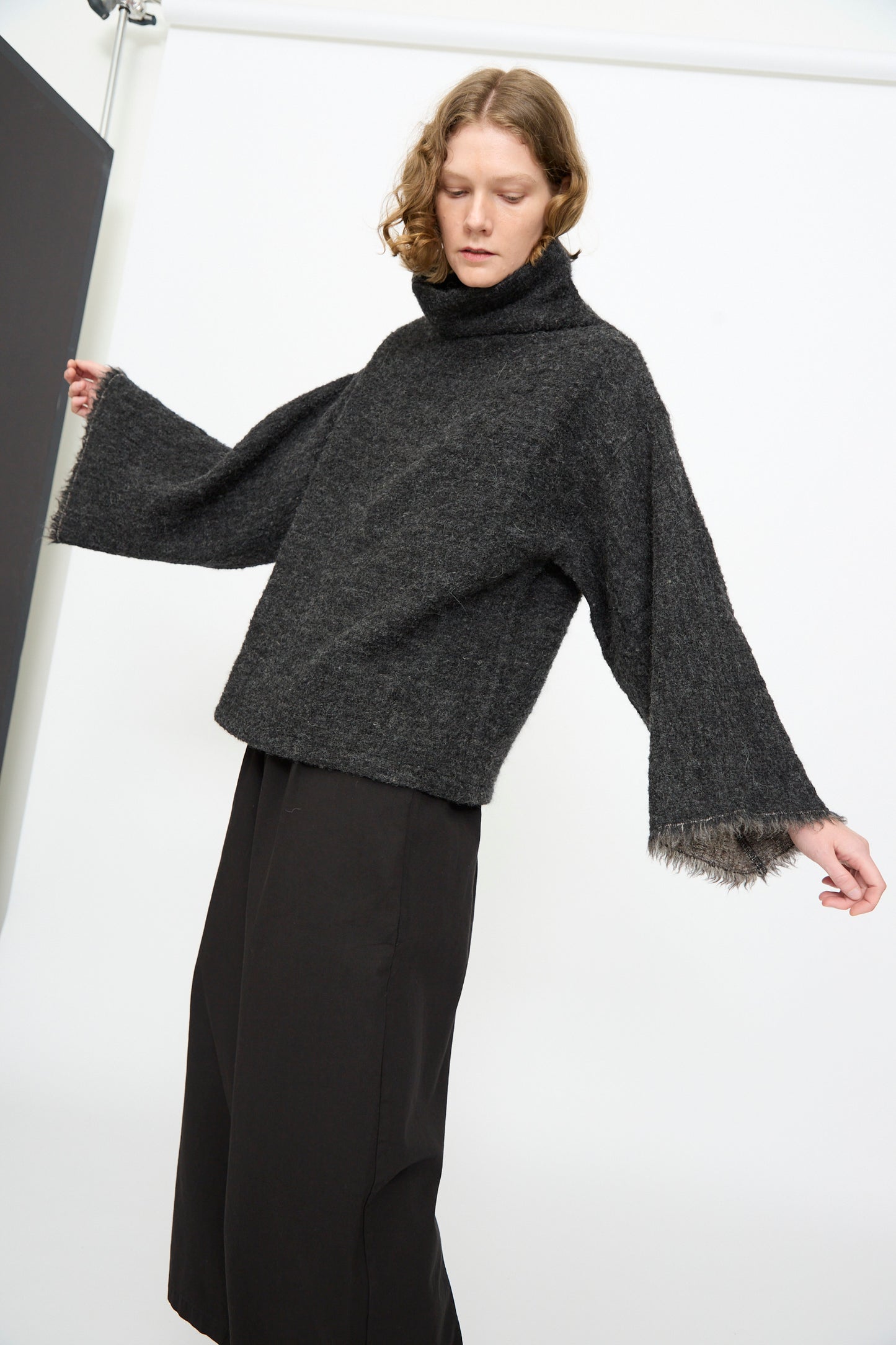 A person wearing the Black Crane Alpaca Tube Top in Dark Grey, styled with wide sleeves and black pants, stands against a plain background.