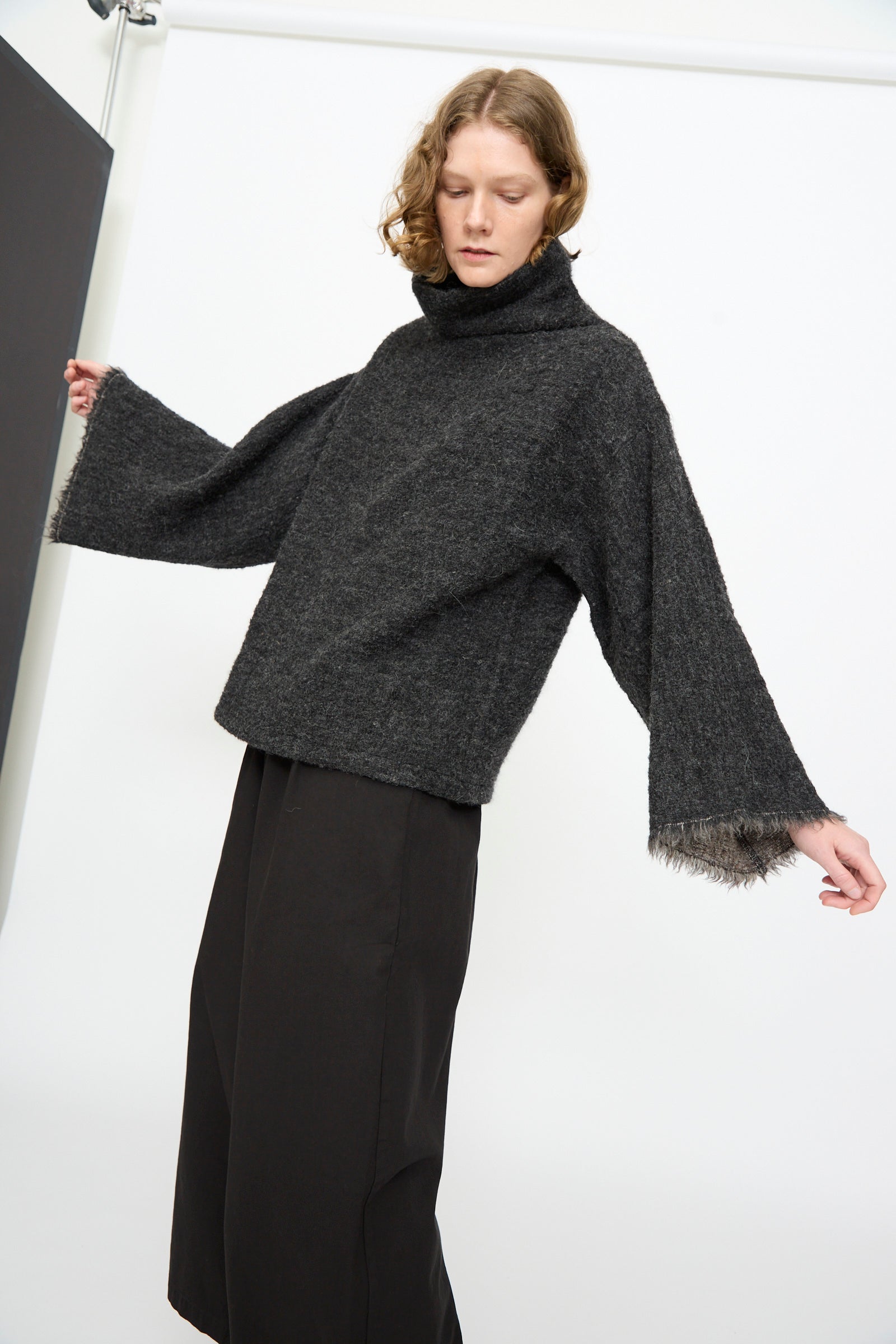 A person wearing the Black Crane Alpaca Tube Top in Dark Grey, styled with wide sleeves and black pants, stands against a plain background.