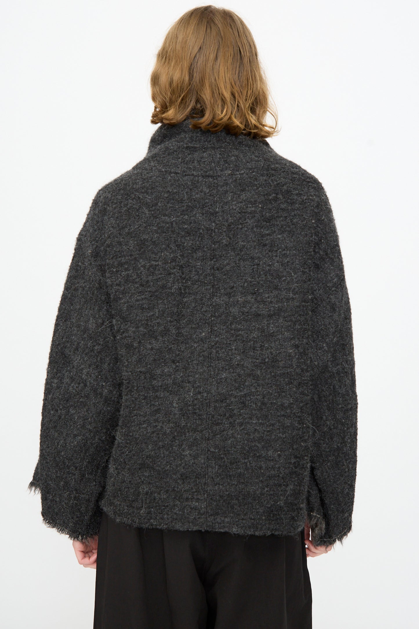A person with shoulder-length light brown hair is seen from behind, wearing a loose, dark gray textured jacket over the Black Crane Alpaca Tube Top in Dark Grey and black pants, set against a plain white background.