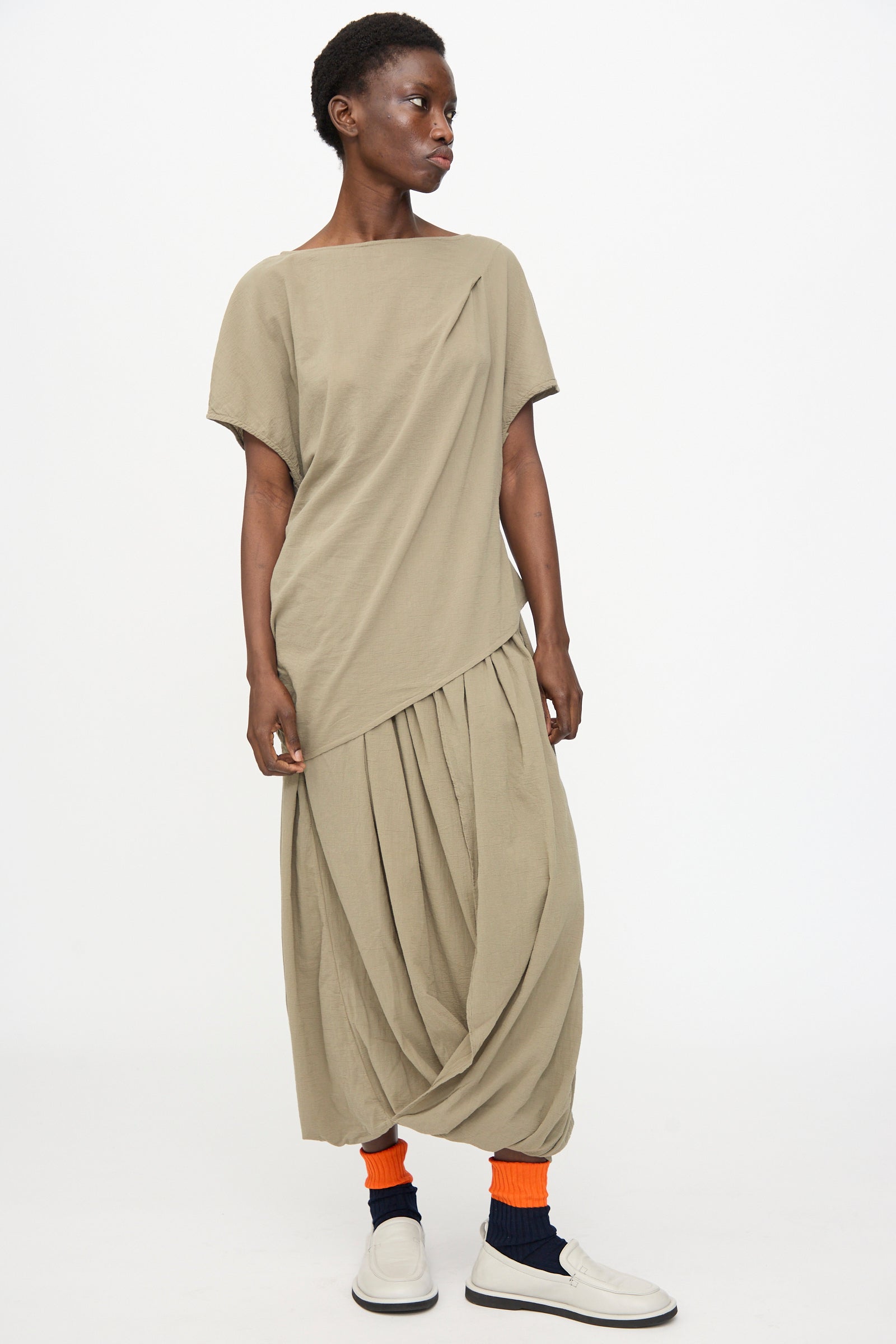 Wearing the Black Crane Asymmetric Top in Cement, a person stands against a plain background, styled with a relaxed skirt, orange socks, navy socks, and white shoes.