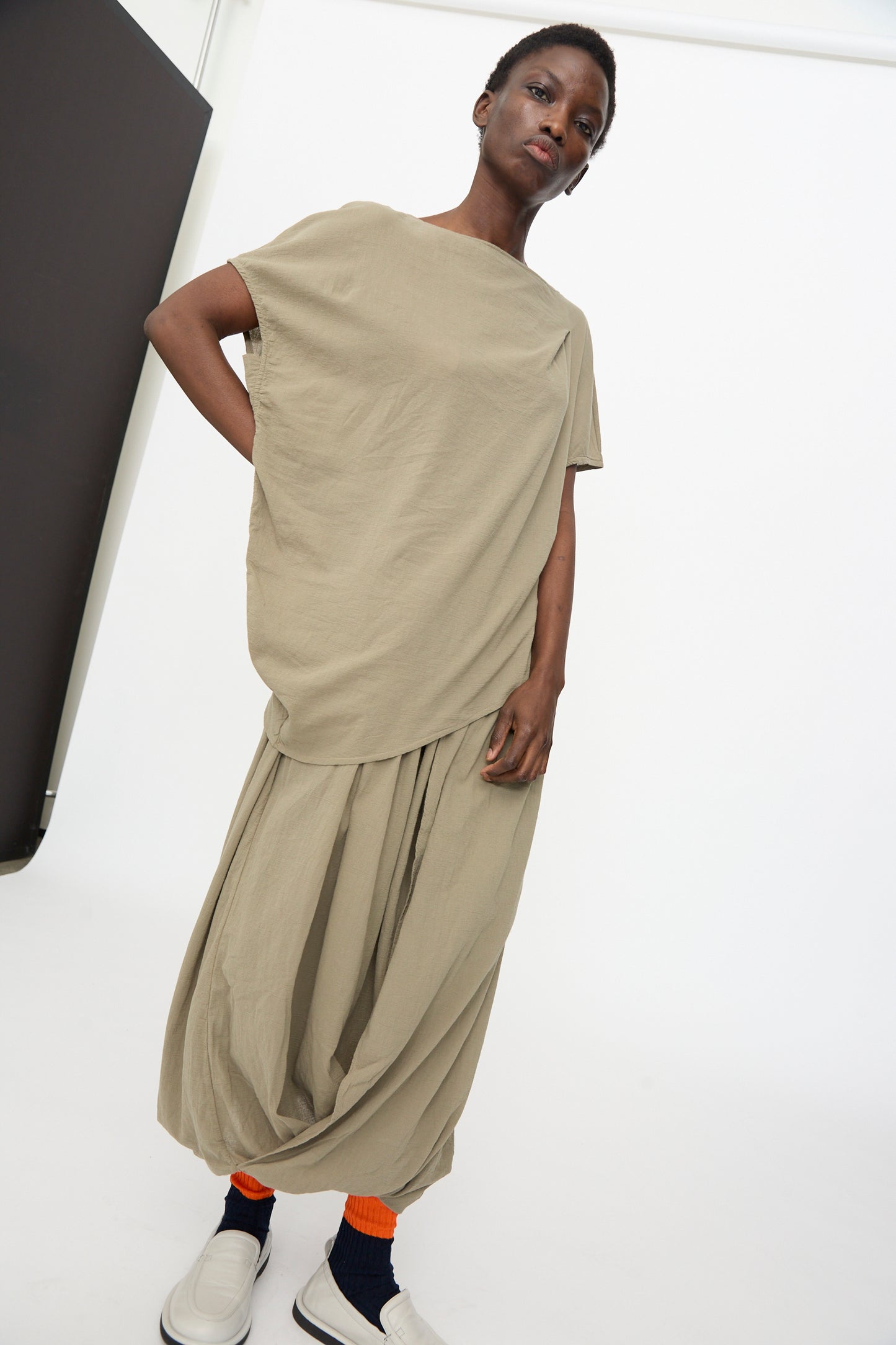 Wearing the Black Crane Asymmetric Top in Cement, paired with a matching draped skirt, bright orange socks, and white slip-on shoes, a person stands against a plain white background.