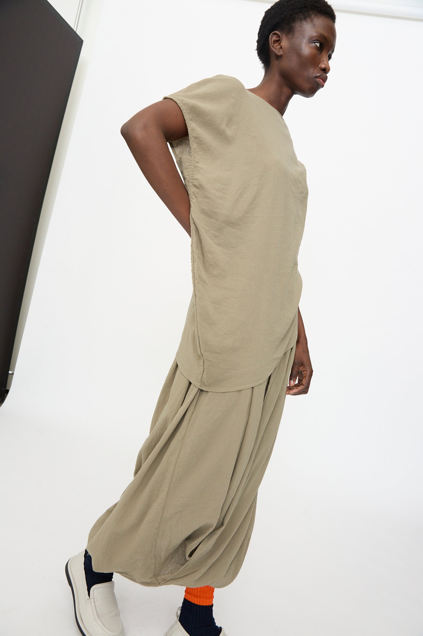 A person stands in a minimalist setting, wearing the Black Crane Asymmetric Top in Cement with matching wide pleated pants, complemented by white shoes and orange socks.