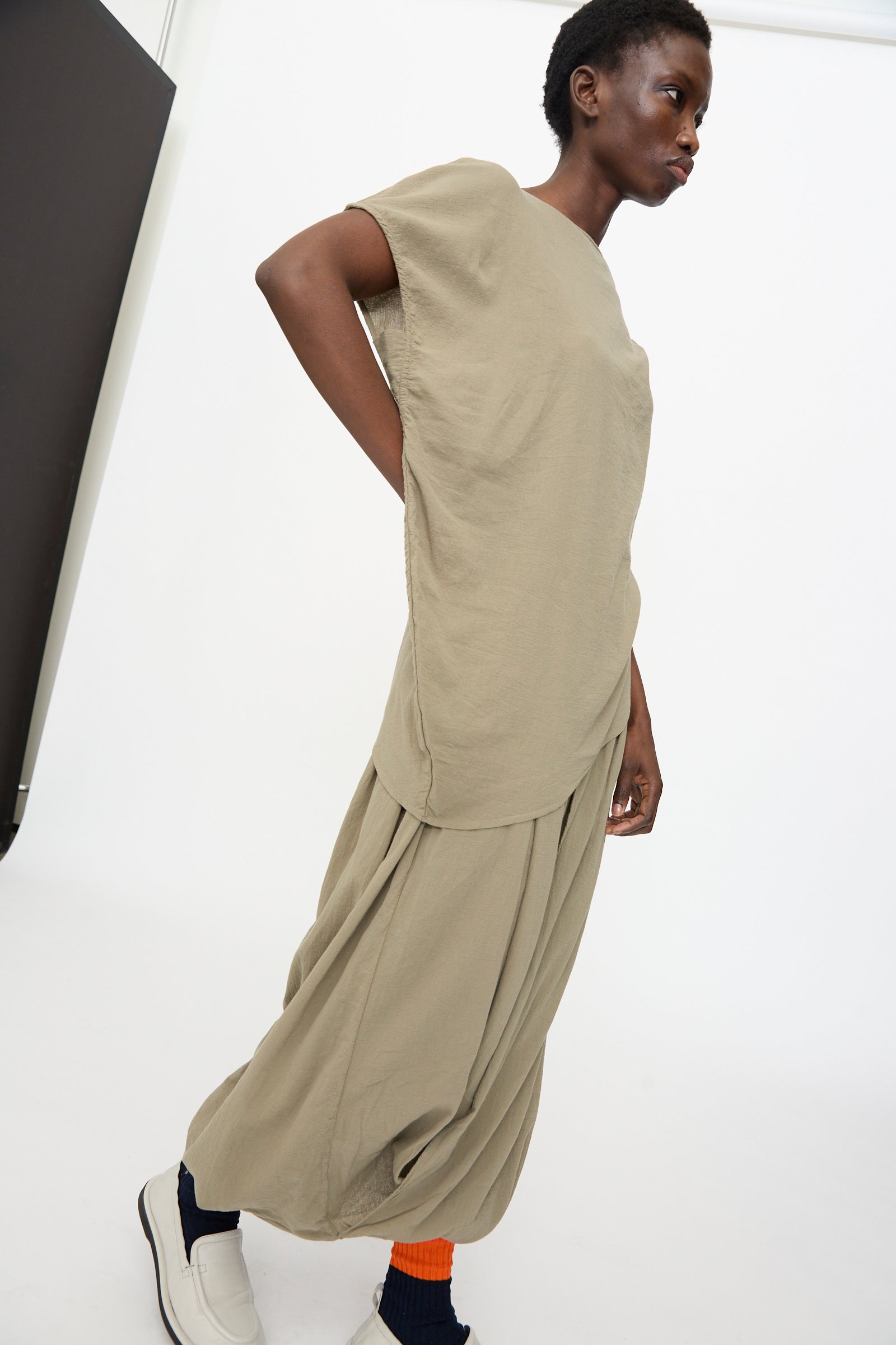 A person stands in a minimalist setting, wearing the Black Crane Asymmetric Top in Cement with matching wide pleated pants, complemented by white shoes and orange socks.