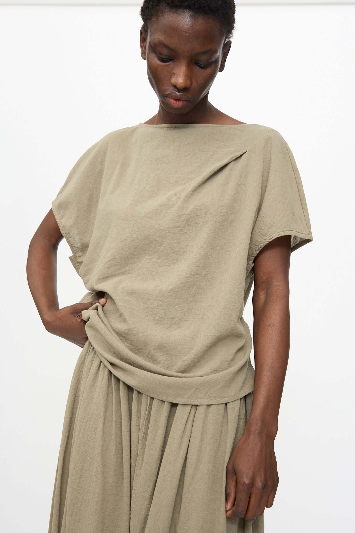 Wearing the Black Crane Asymmetric Top in Cement, a person stands against a plain white background, looking downward with one hand on their waist.