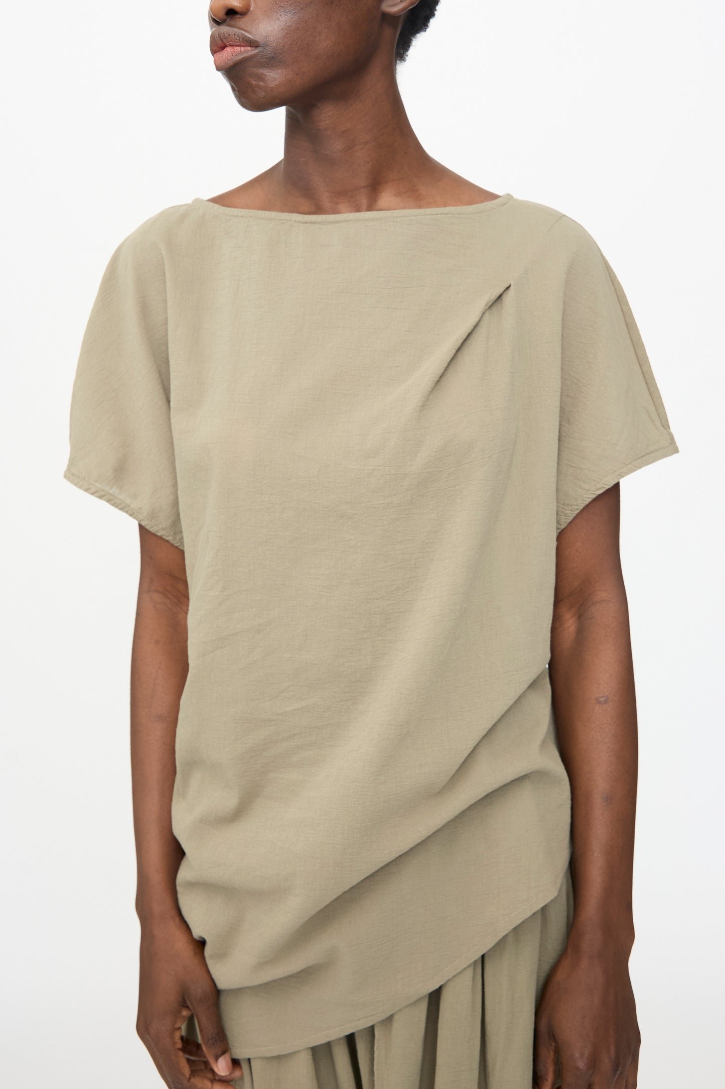 A person wears the Black Crane Asymmetric Top in Cement, a relaxed fit short sleeve top with pleat detail near the neckline, paired with a matching skirt, standing against a plain background.