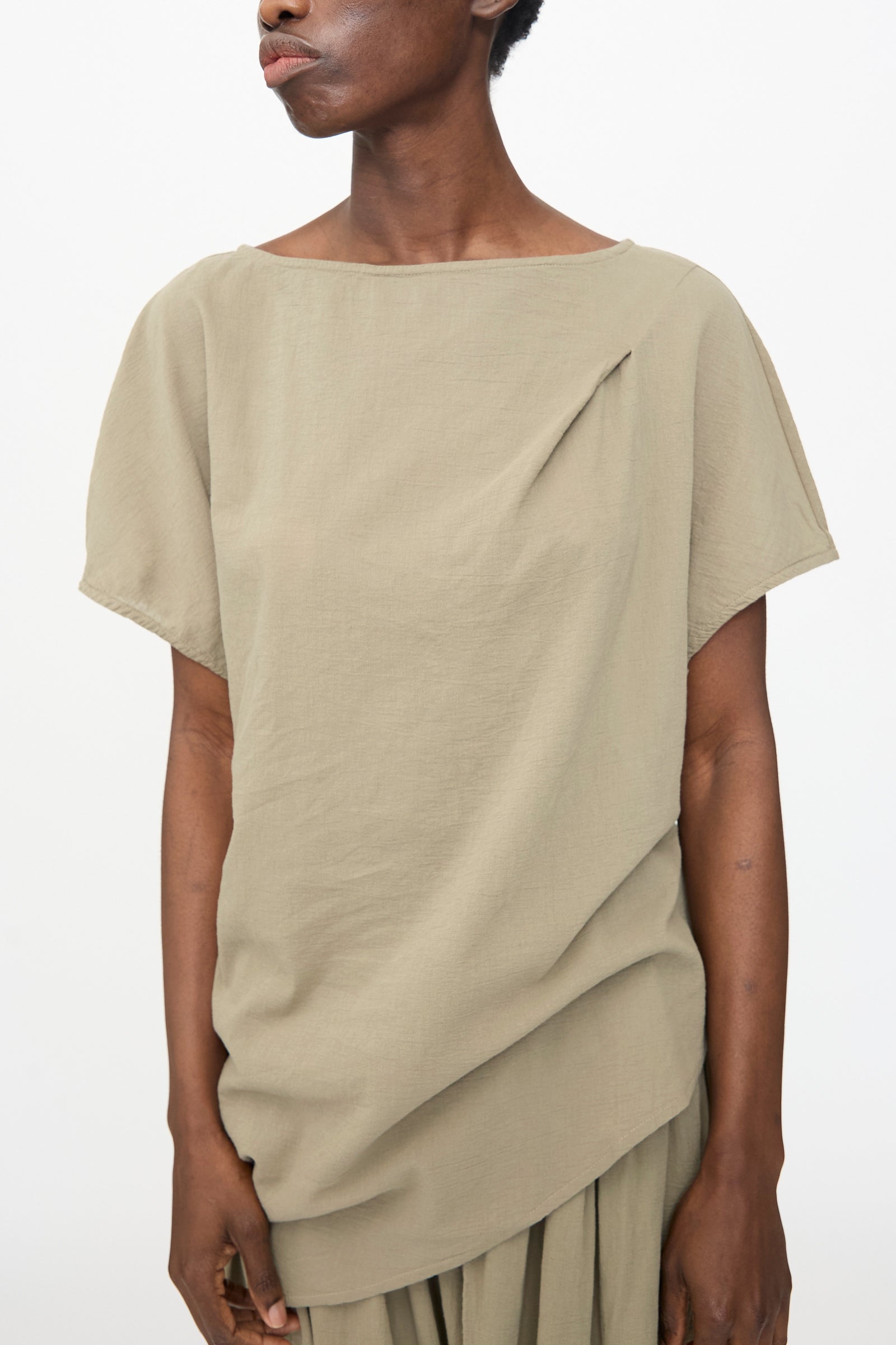 A person wears the Black Crane Asymmetric Top in Cement, a relaxed fit short sleeve top with pleat detail near the neckline, paired with a matching skirt, standing against a plain background.