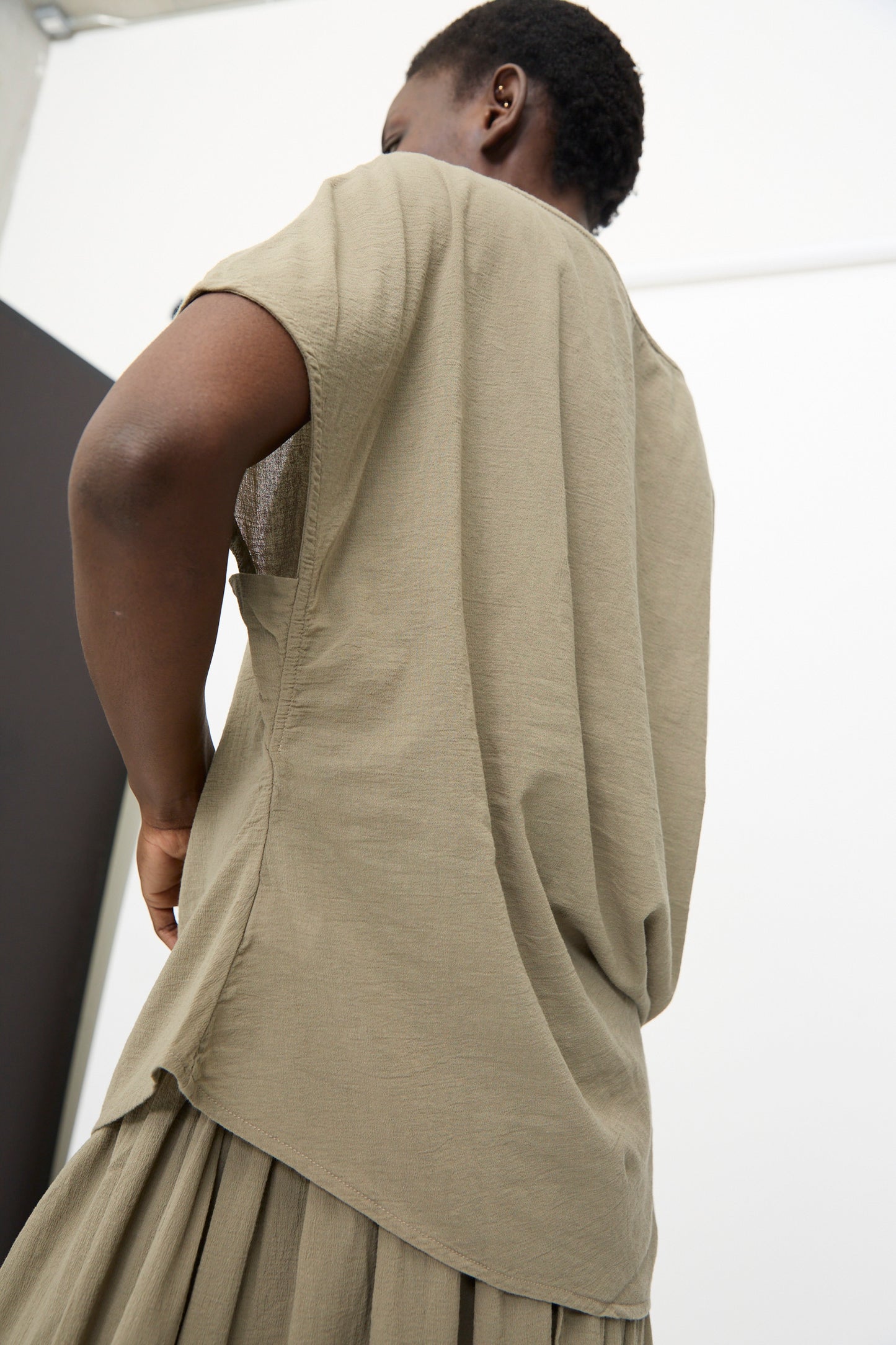 Person wearing the Black Crane Asymmetric Top in Cement, a relaxed fit short sleeve cotton gauze top with pleated detail at the bottom, photographed from the back against a plain indoor background.
