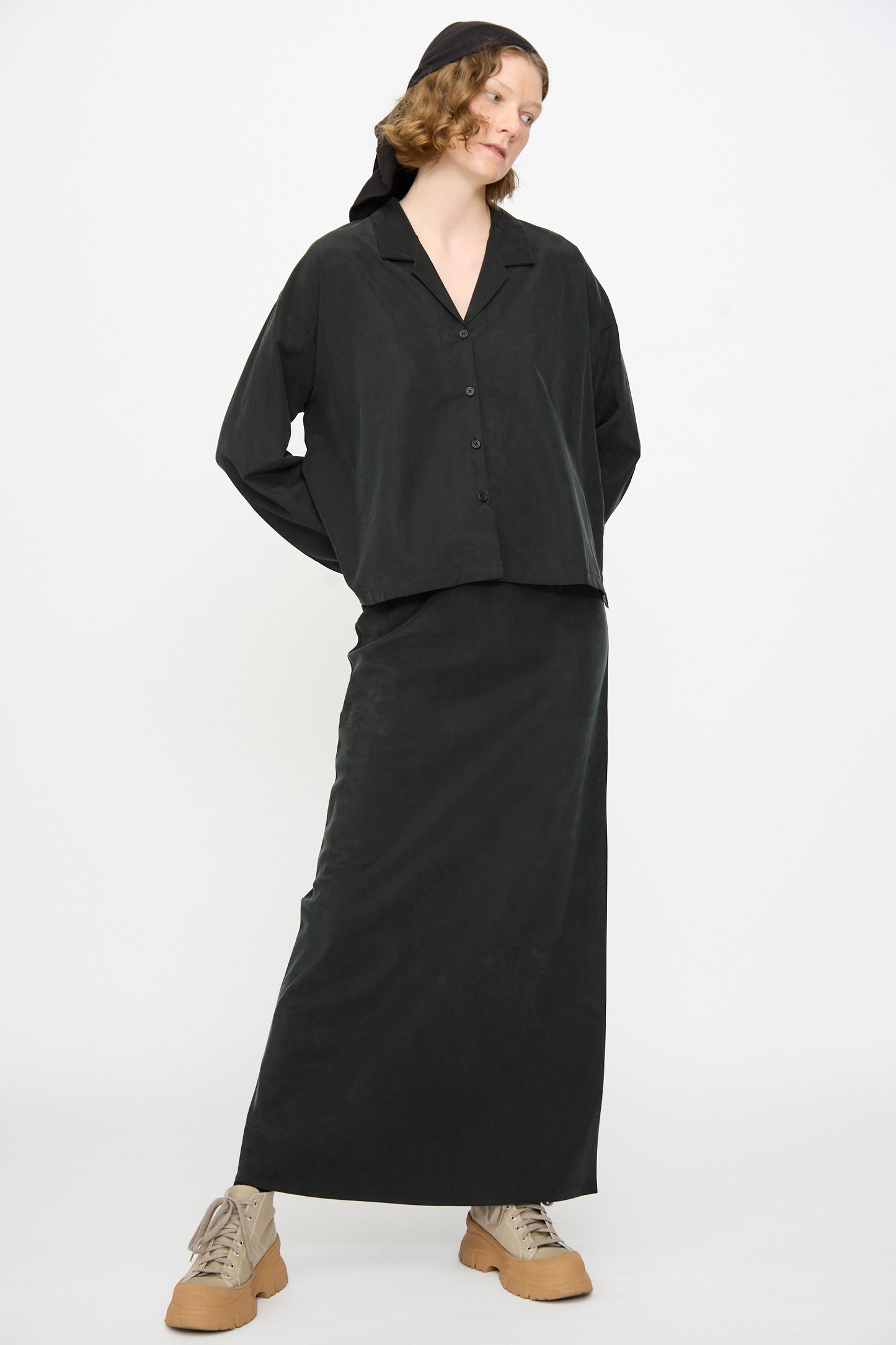A person wearing a black button-up shirt, the Black Crane Bias Skirt in Black, a black headscarf, and beige lace-up shoes stands against a plain white background.