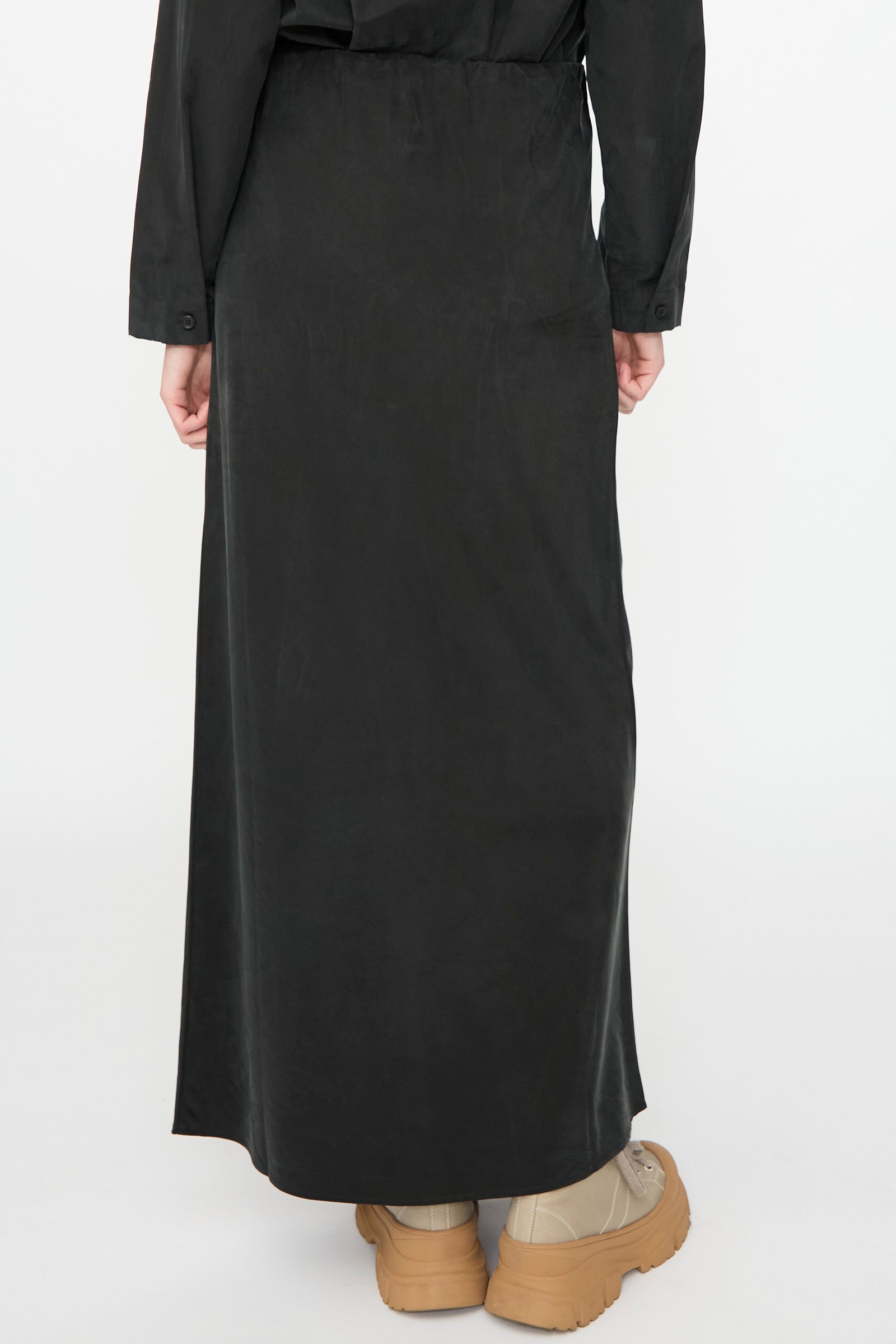 A person is shown from the back against a plain white background, wearing the Black Crane Bias Skirt in Black—a long maxi skirt crafted from a soft cotton cupro blend with an elastic waistband for added comfort—paired with tan sneakers.