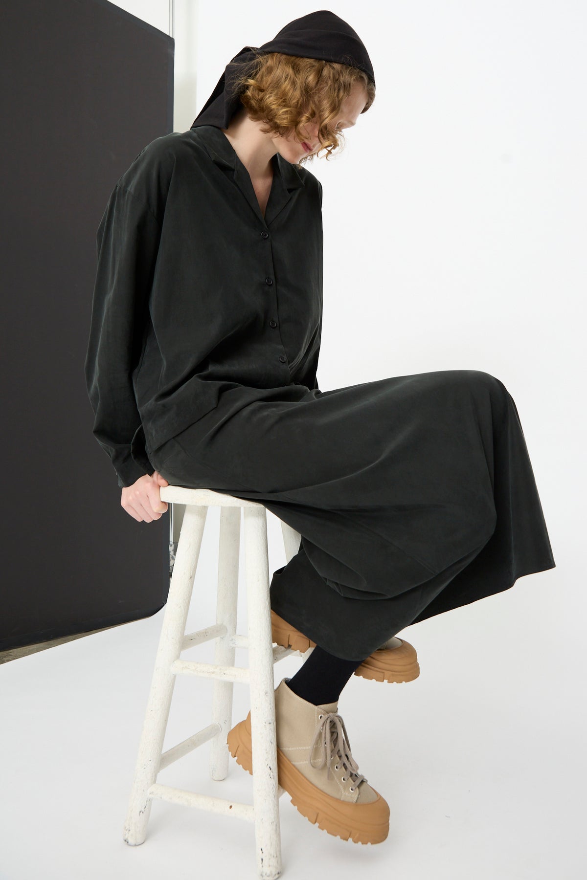 A person in loose black clothing and tan platform shoes sits on a white stool against a plain background, looking downward with their head slightly turned and wearing a black headscarf. Their outfit features the Black Crane Bias Skirt in Black—an elegant maxi skirt crafted from a soft cotton cupro blend with an elastic waistband for effortless comfort.