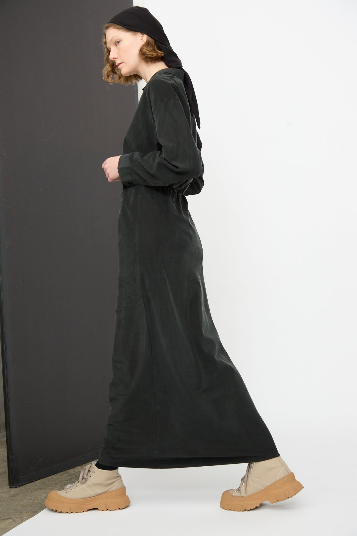 A person wearing the Black Crane Bias Skirt in Black, paired with tan boots and a black headscarf, walks indoors against a background split into black and white.