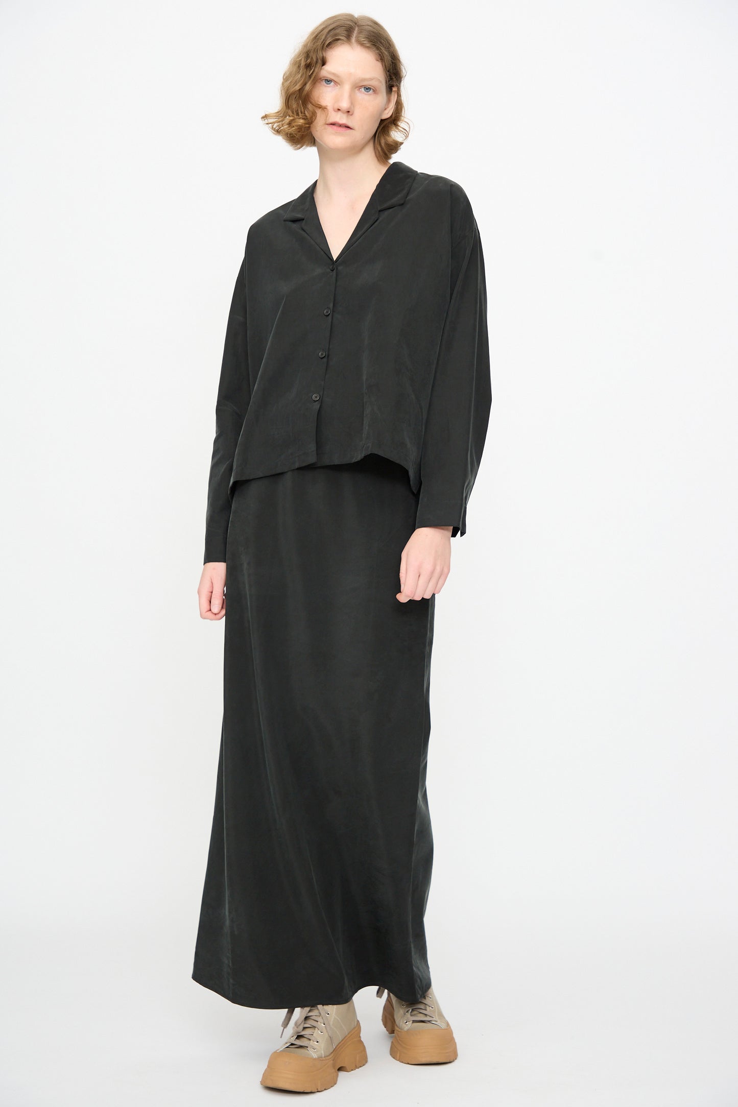 A person wearing the Black Crane Bias Skirt in Black—a loose-fitting black maxi skirt with an elastic waistband, crafted from a soft cotton cupro blend—paired with a matching black button-up shirt and beige lace-up shoes, stands against a plain white background.