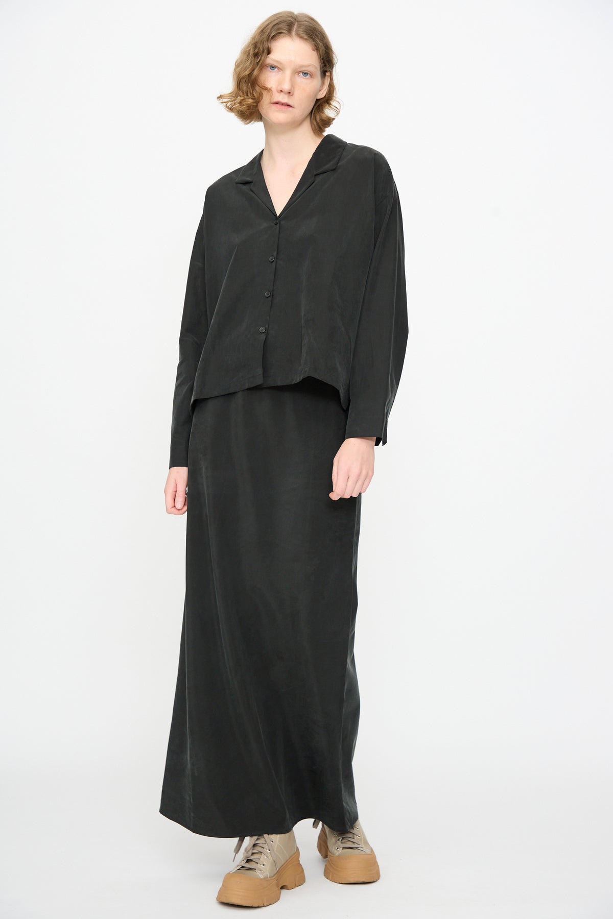 A person wearing the Black Crane Bias Skirt in Black—a loose-fitting black maxi skirt with an elastic waistband, crafted from a soft cotton cupro blend—paired with a matching black button-up shirt and beige lace-up shoes, stands against a plain white background.
