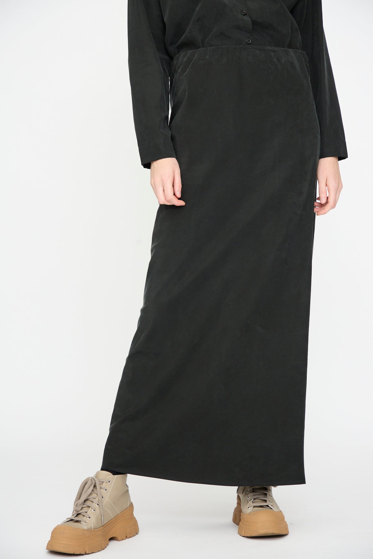 A person stands against a plain white background wearing the Black Crane Bias Skirt in Black—a long maxi skirt featuring an elastic waistband and crafted from a soft cotton cupro blend—paired with a black long-sleeve top and beige lace-up boots.