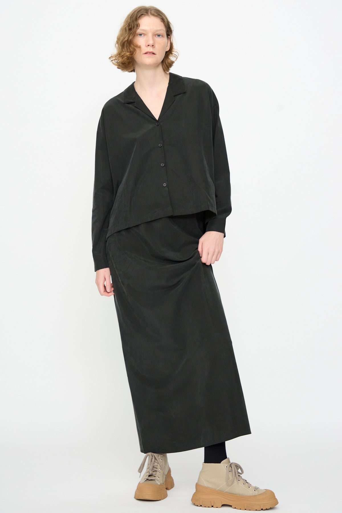 A person is wearing the Black Crane Bias Skirt in Black—a loose, maxi-length style with an elastic waistband crafted from a soft cotton cupro blend—paired with a black button-up shirt, black leggings, and tan lace-up shoes, standing against a plain white background.