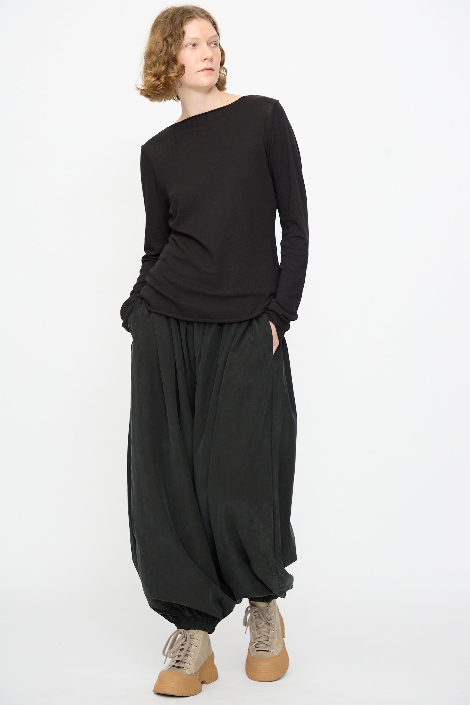 A person stands against a white background, wearing the Black Crane Boat Neck in Black— a relaxed fit, long-sleeve top—paired with loose black pants and tan lace-up shoes, hands in pockets and head slightly turned.