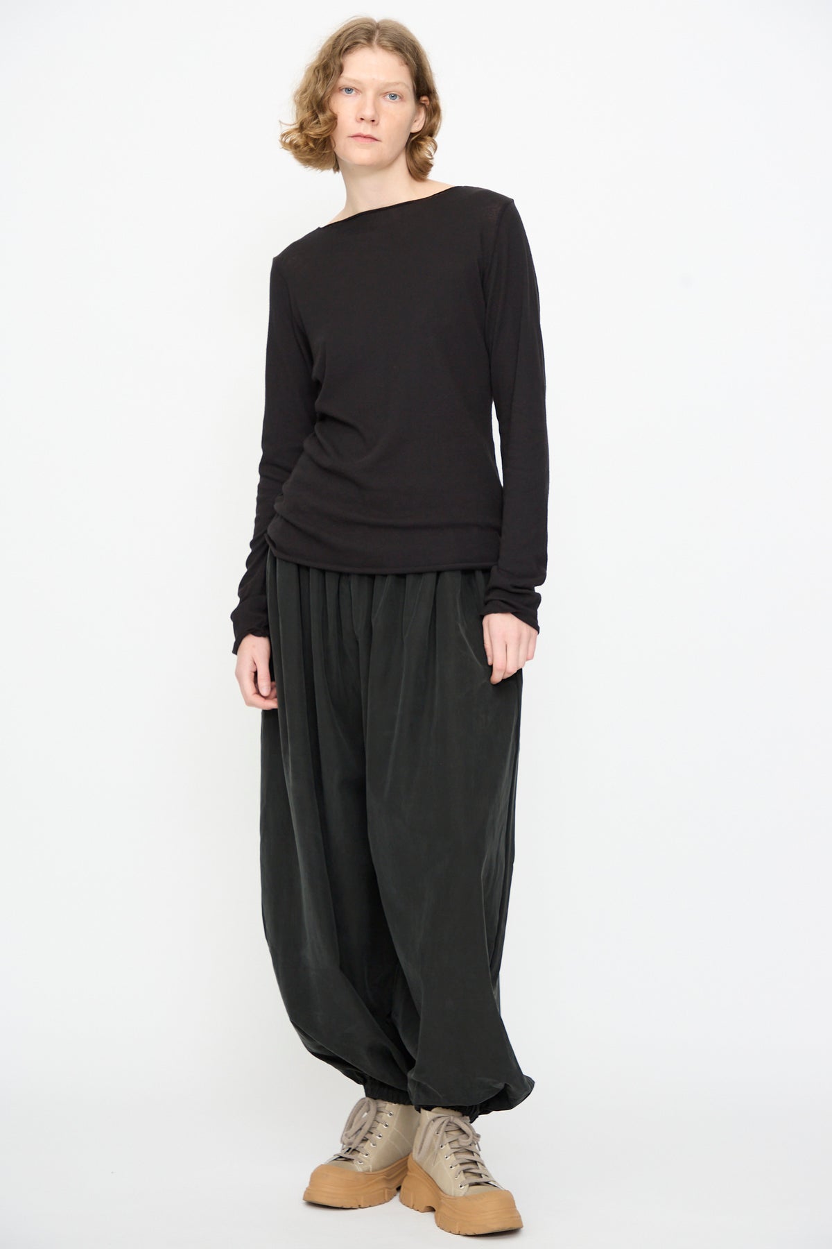 A person is standing against a plain white background, wearing the Black Crane Boat Neck in Black—a relaxed fit long sleeve top—paired with loose dark pants and beige lace-up shoes.
