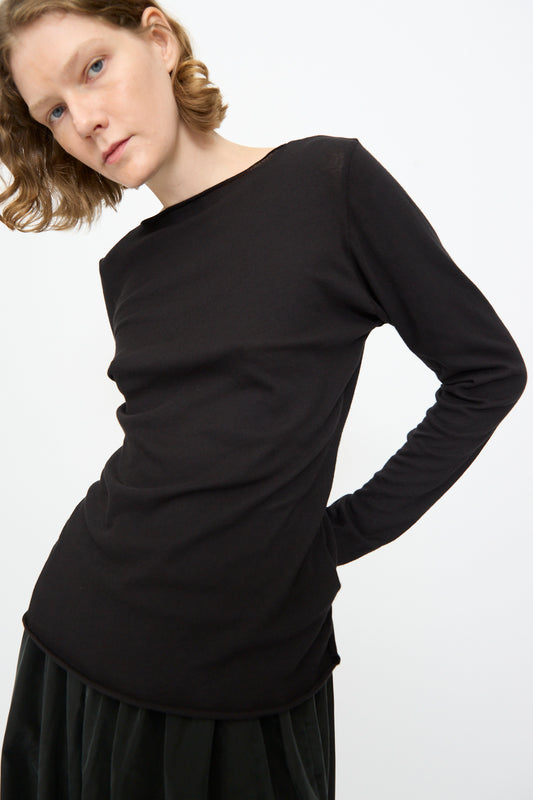 A person with short, wavy hair wears the Black Crane Boat Neck in Black—a relaxed-fit long sleeve top—paired with a dark skirt, standing against a plain white background and looking slightly downward with a neutral expression.