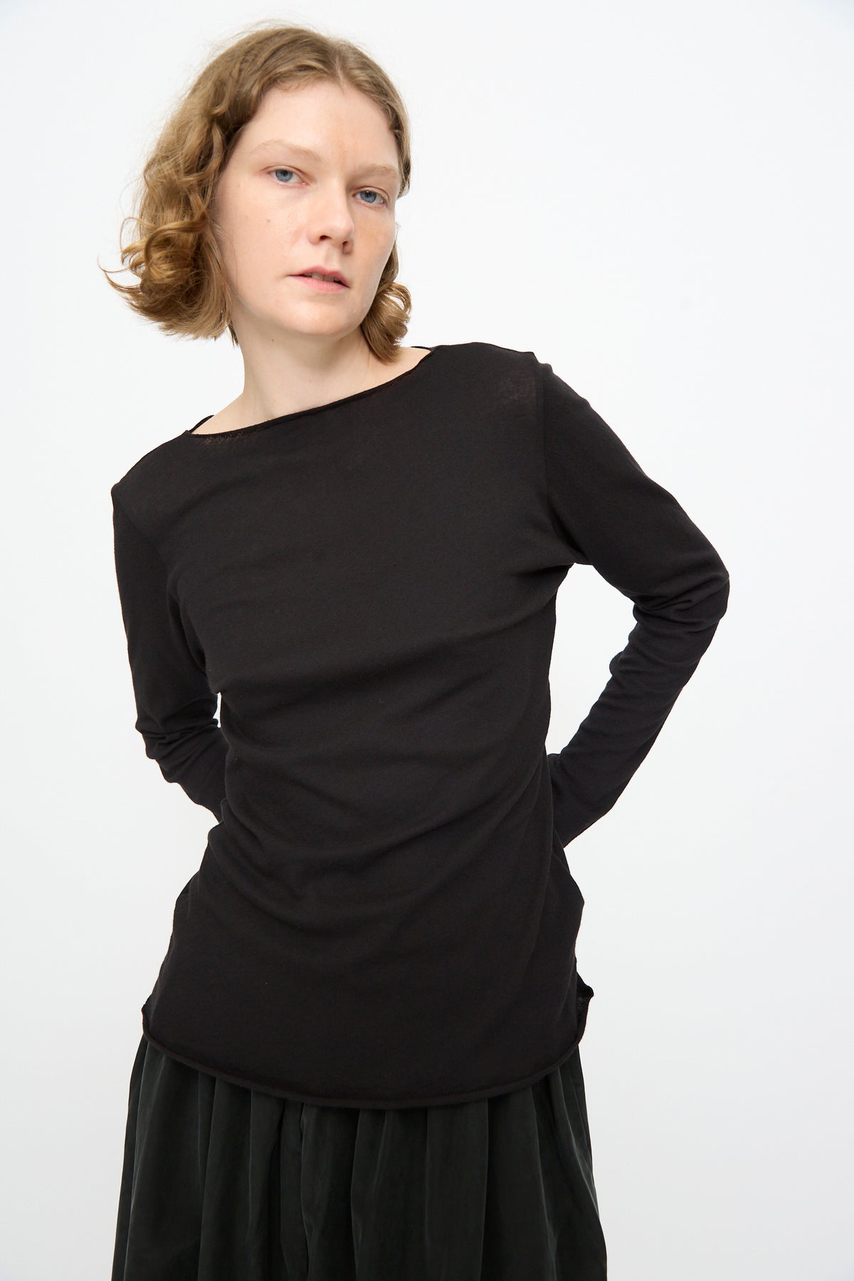 A person with short, wavy hair wears the Black Crane Boat Neck in Black, a relaxed fit long sleeve cotton-cashmere top, paired with a dark skirt. They stand against a plain white background with hands behind their back.