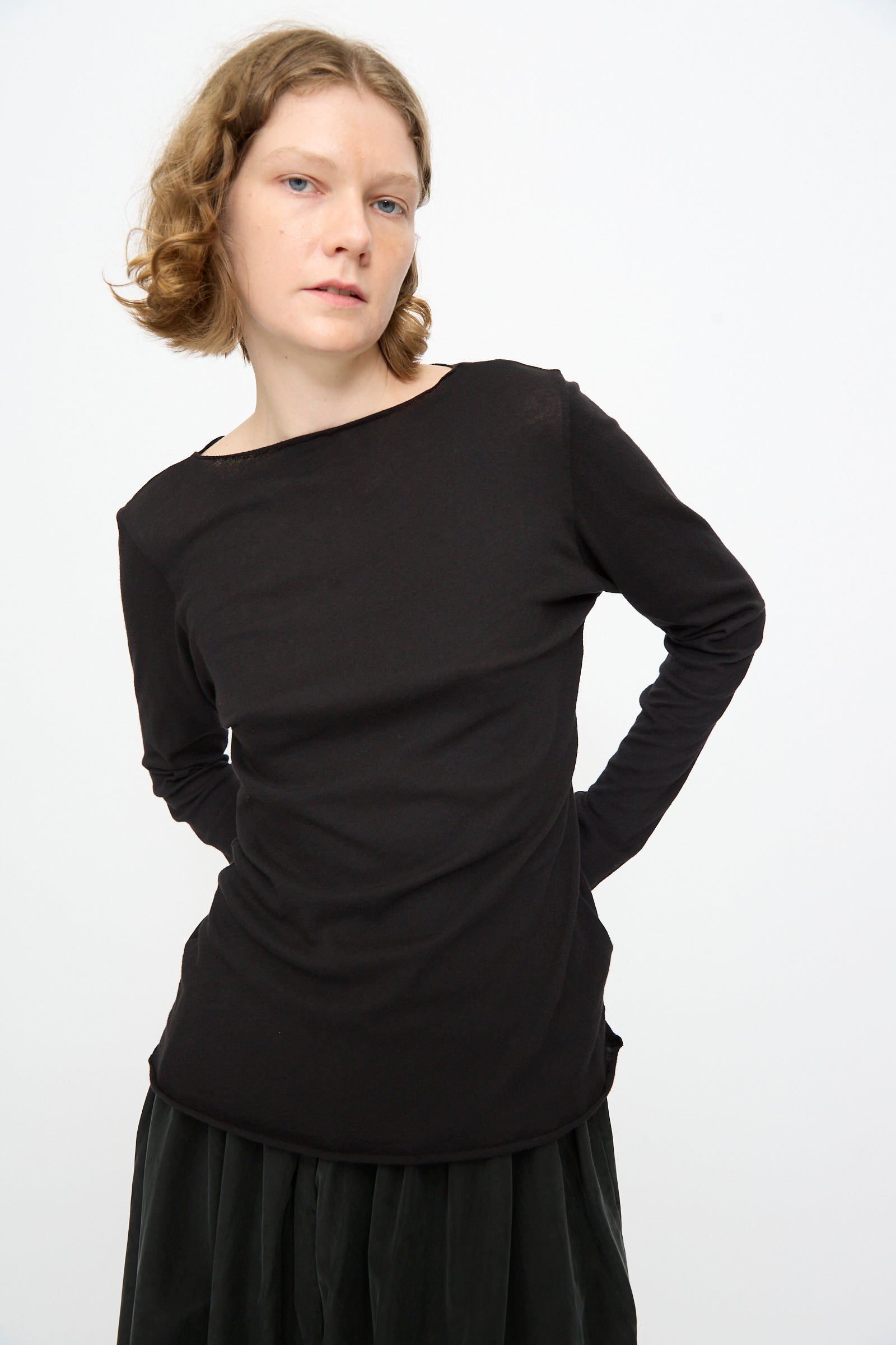 A person with short, wavy hair wears the Black Crane Boat Neck in Black, a relaxed fit long sleeve cotton-cashmere top, paired with a dark skirt. They stand against a plain white background with hands behind their back.