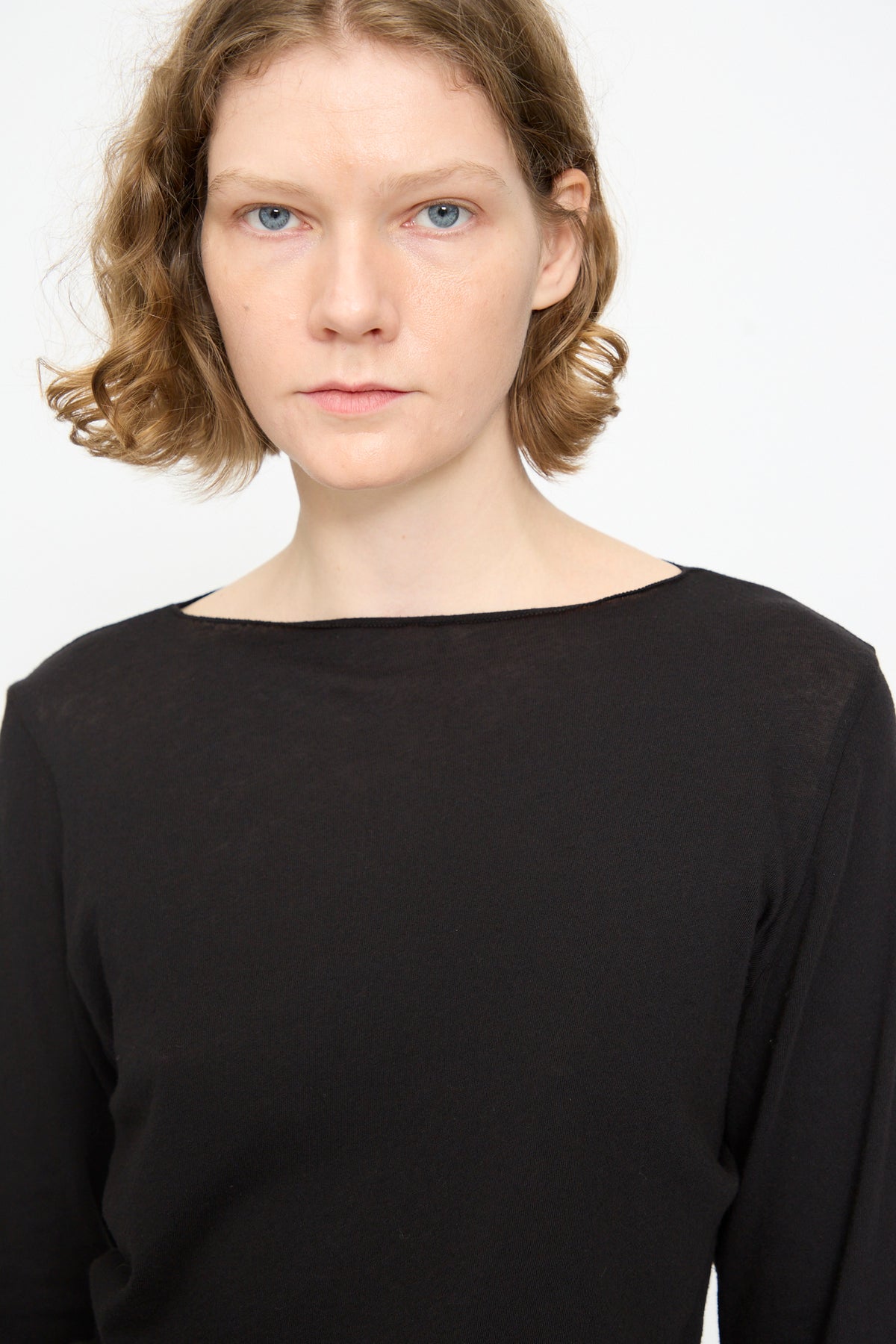 A person with short, wavy light brown hair is wearing the Black Crane Boat Neck in Black, a relaxed fit long sleeve top, and is standing against a white background with a neutral expression.