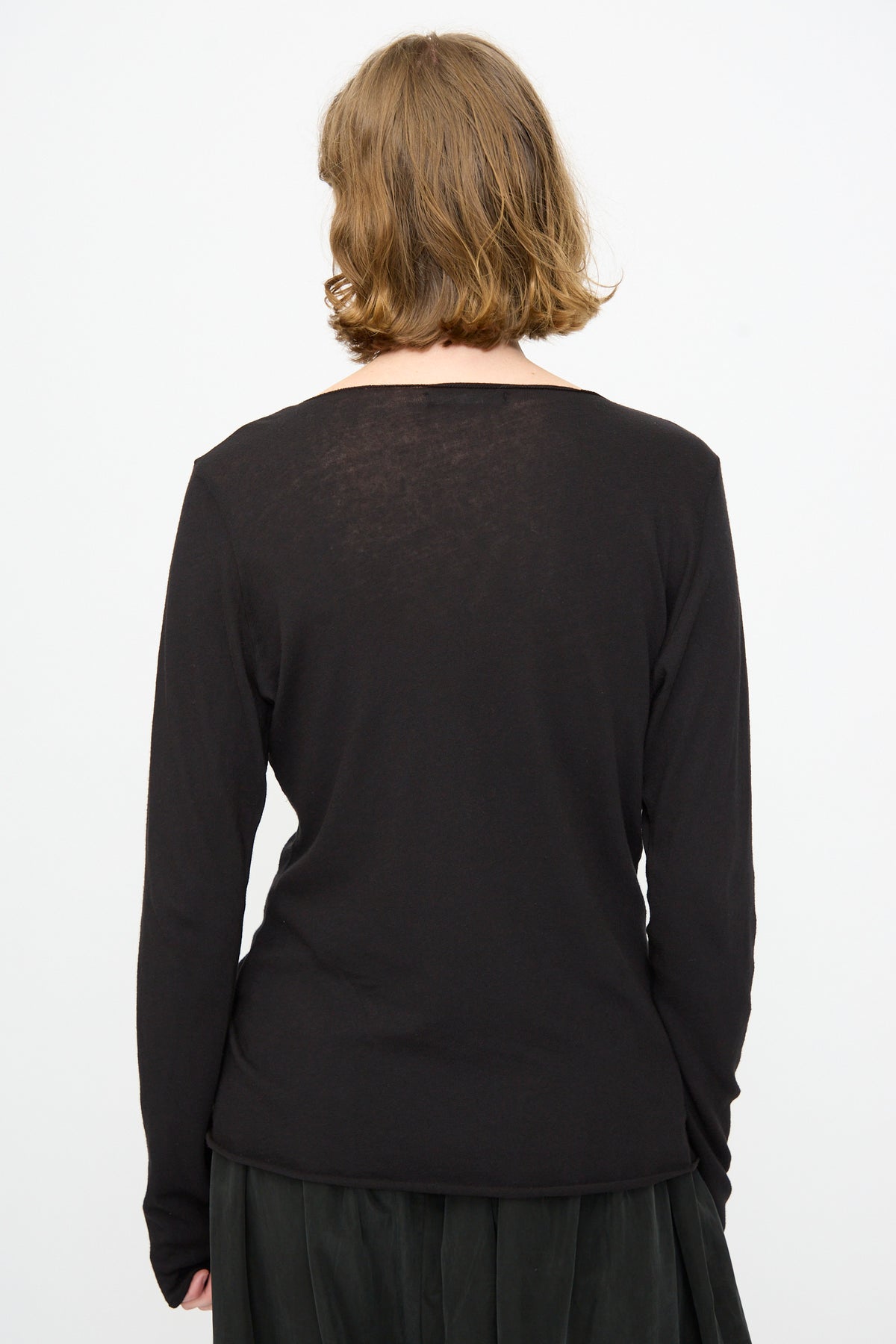 A person with short, light brown hair is shown from the back, wearing the Black Crane Boat Neck in Black—a relaxed fit long sleeve top—paired with a dark skirt, set against a plain white background.