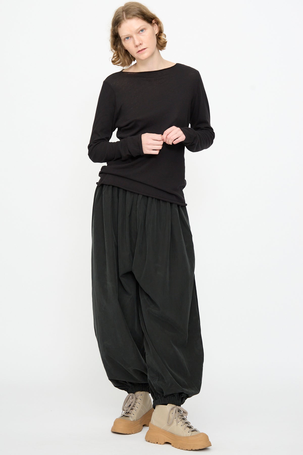 A person stands against a white background wearing the Black Crane Boat Neck in Black, relaxed-fit harem pants gathered at the ankles, and beige lace-up shoes.