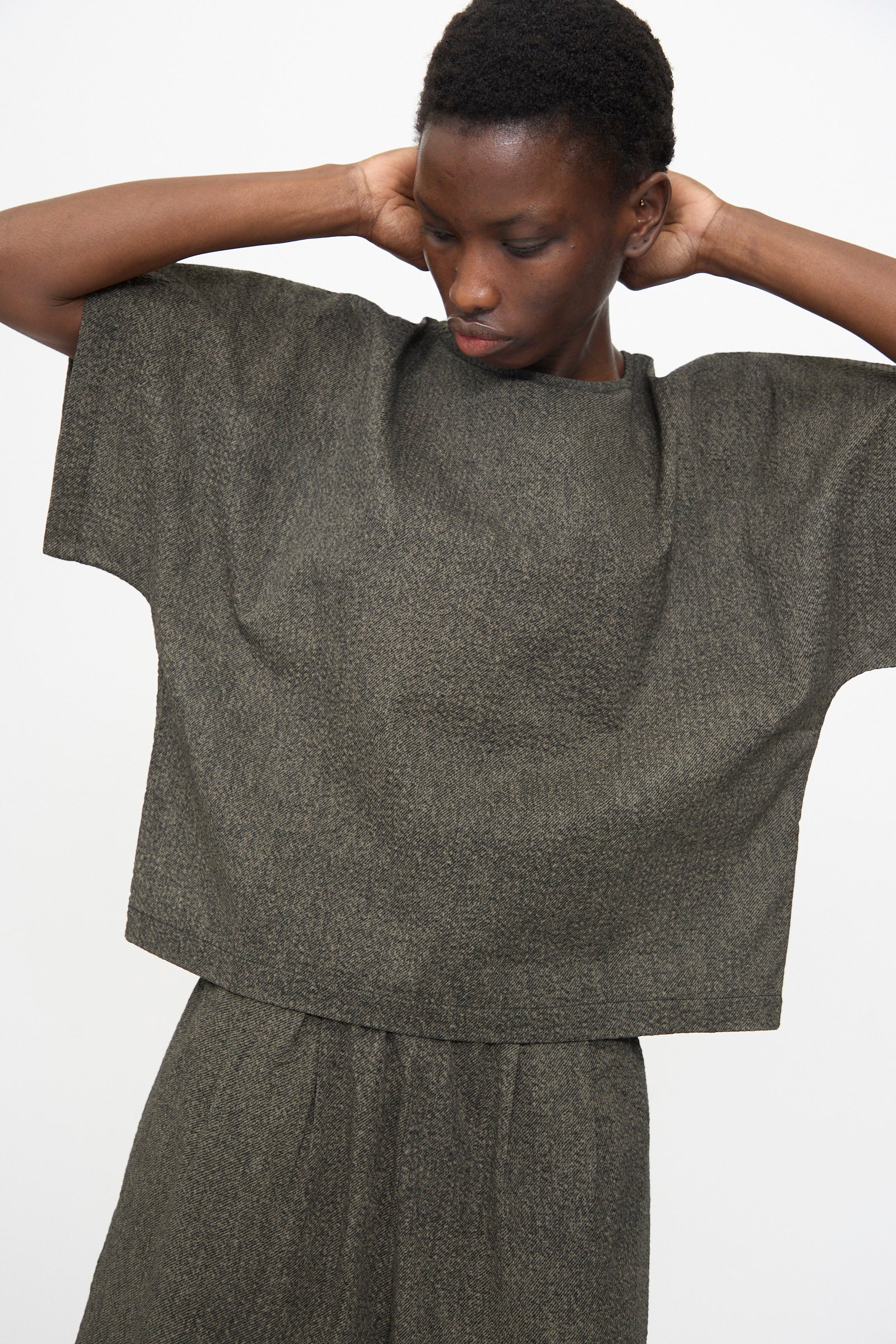 A person stands against a plain background, wearing the Black Crane Box Top with Micro Wave Texture in Steel, paired with matching trousers, hands behind their neck.