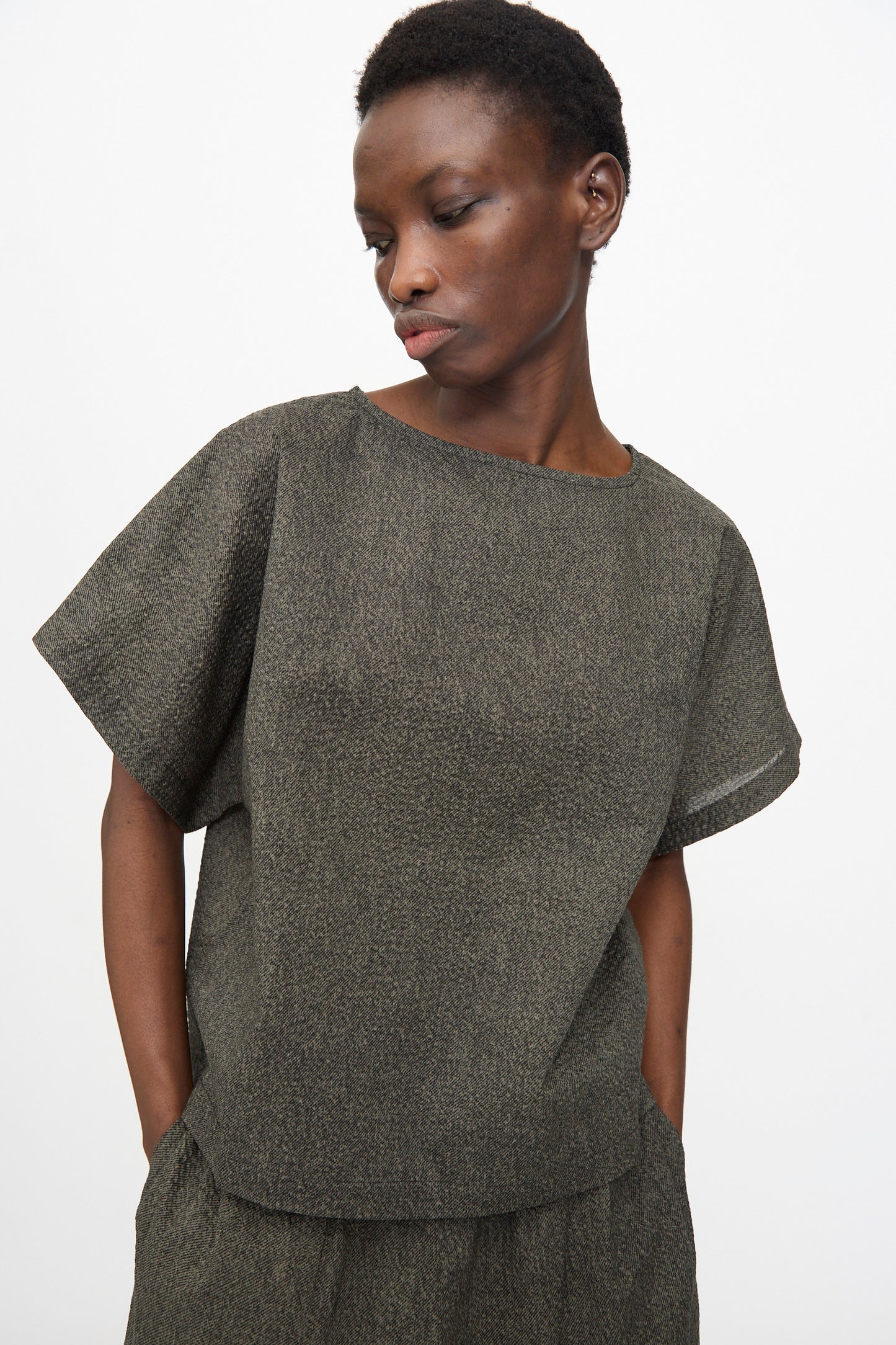 Wearing the Black Crane Box Top with Micro Wave Texture in Steel, the person stands against a plain white background, looking down and to the side in this relaxed fit Japanese cotton shirt.