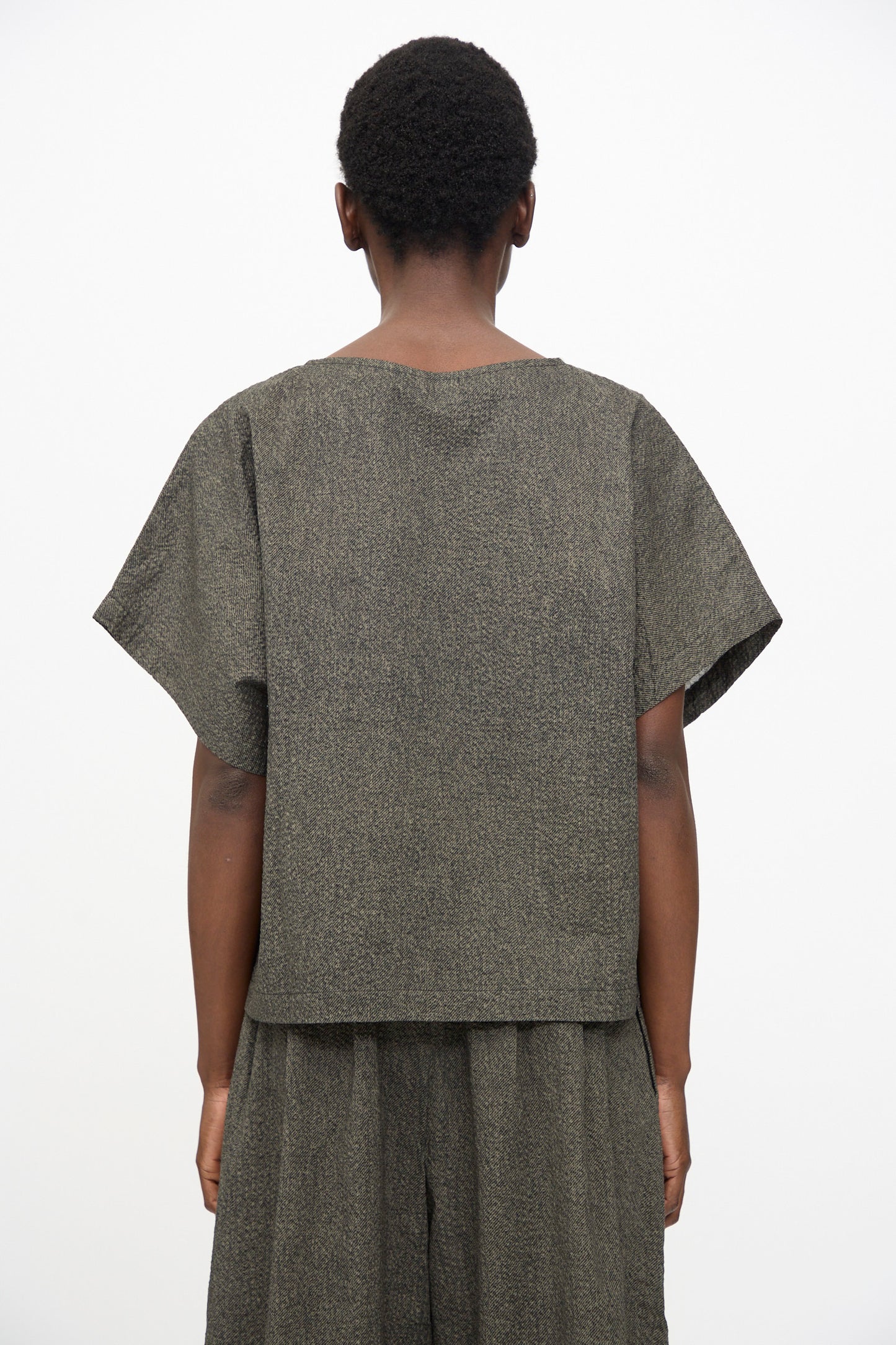 A person stands with their back to the camera, wearing Black Crane’s Box Top with Micro Wave Texture in Steel—a relaxed fit, boxy short sleeve top paired with matching wide-leg pants in a muted dark tone.