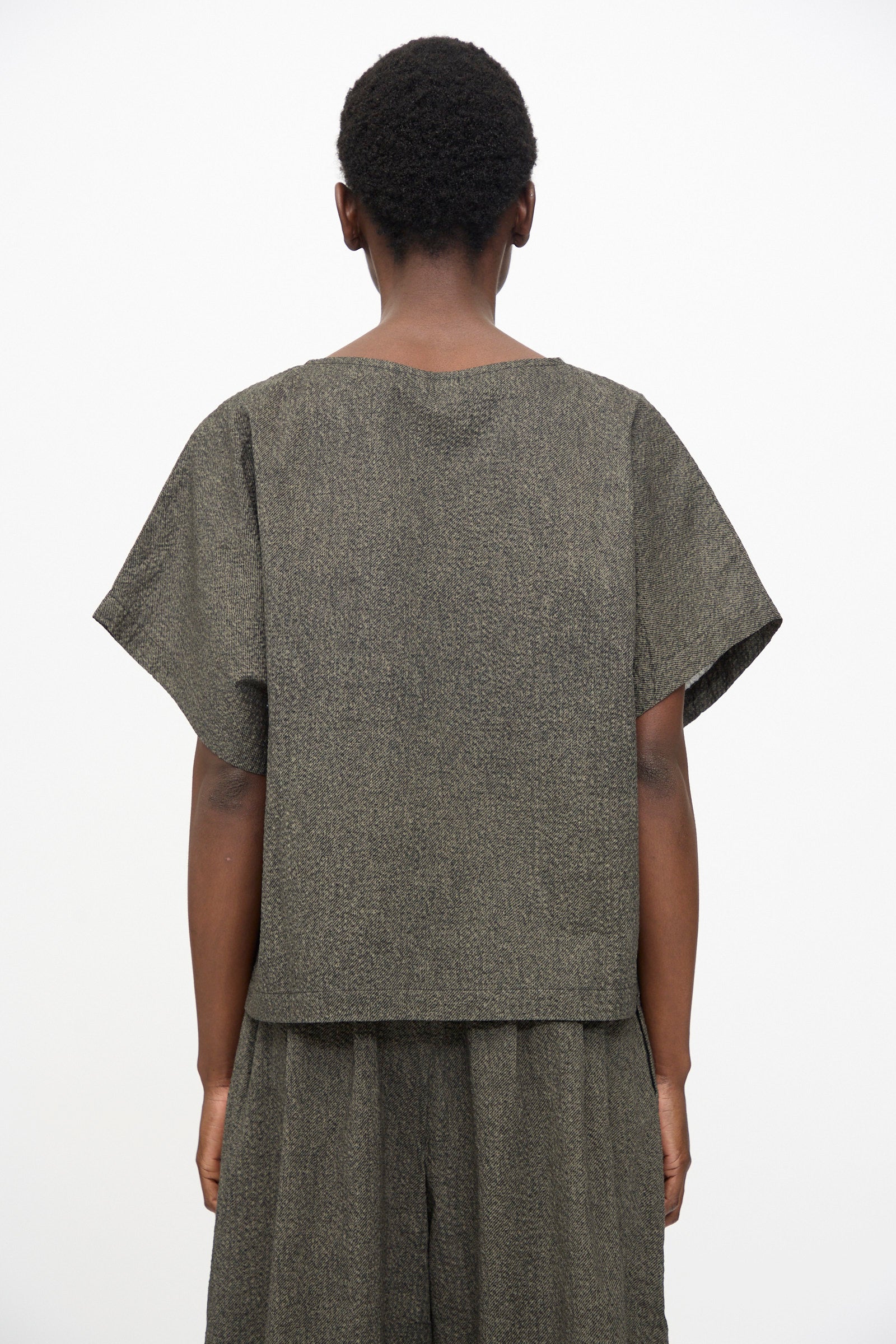A person stands with their back to the camera, wearing Black Crane’s Box Top with Micro Wave Texture in Steel—a relaxed fit, boxy short sleeve top paired with matching wide-leg pants in a muted dark tone.