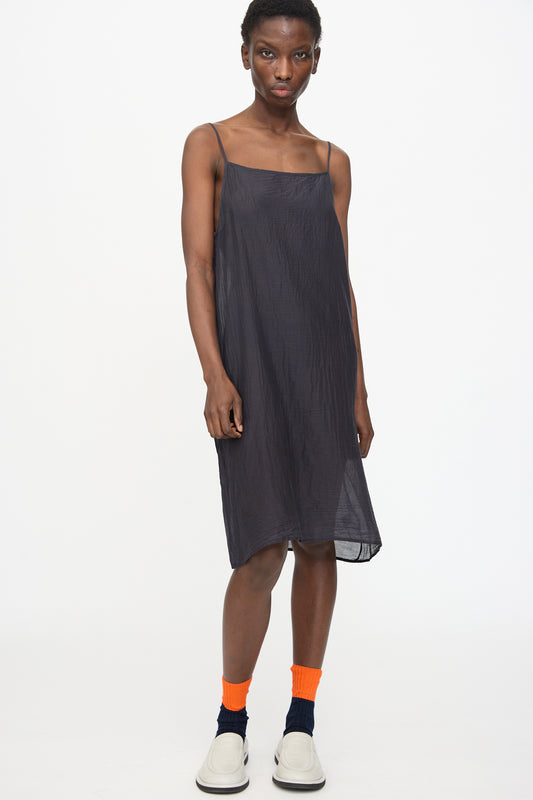 A person stands against a white background, wearing the Black Crane Cami Dress in Faded Black with adjustable straps, paired with orange and black socks and white slip-on shoes.