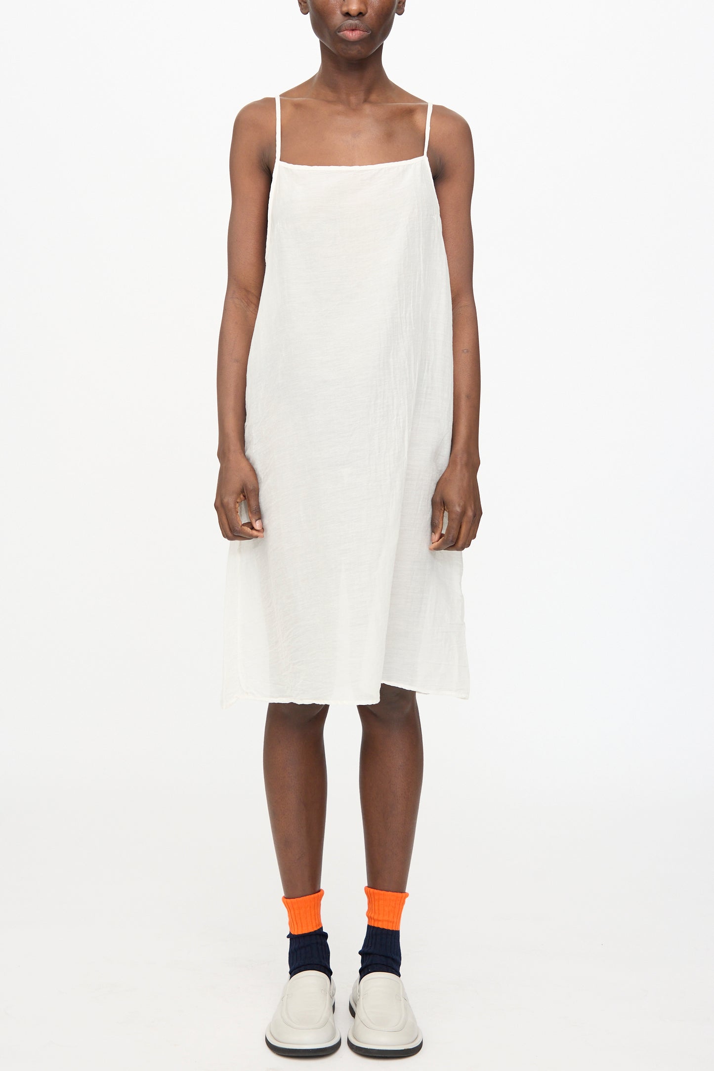 A person stands against a white background wearing the Black Crane Cami Dress in White, paired with orange and navy socks and white shoes.