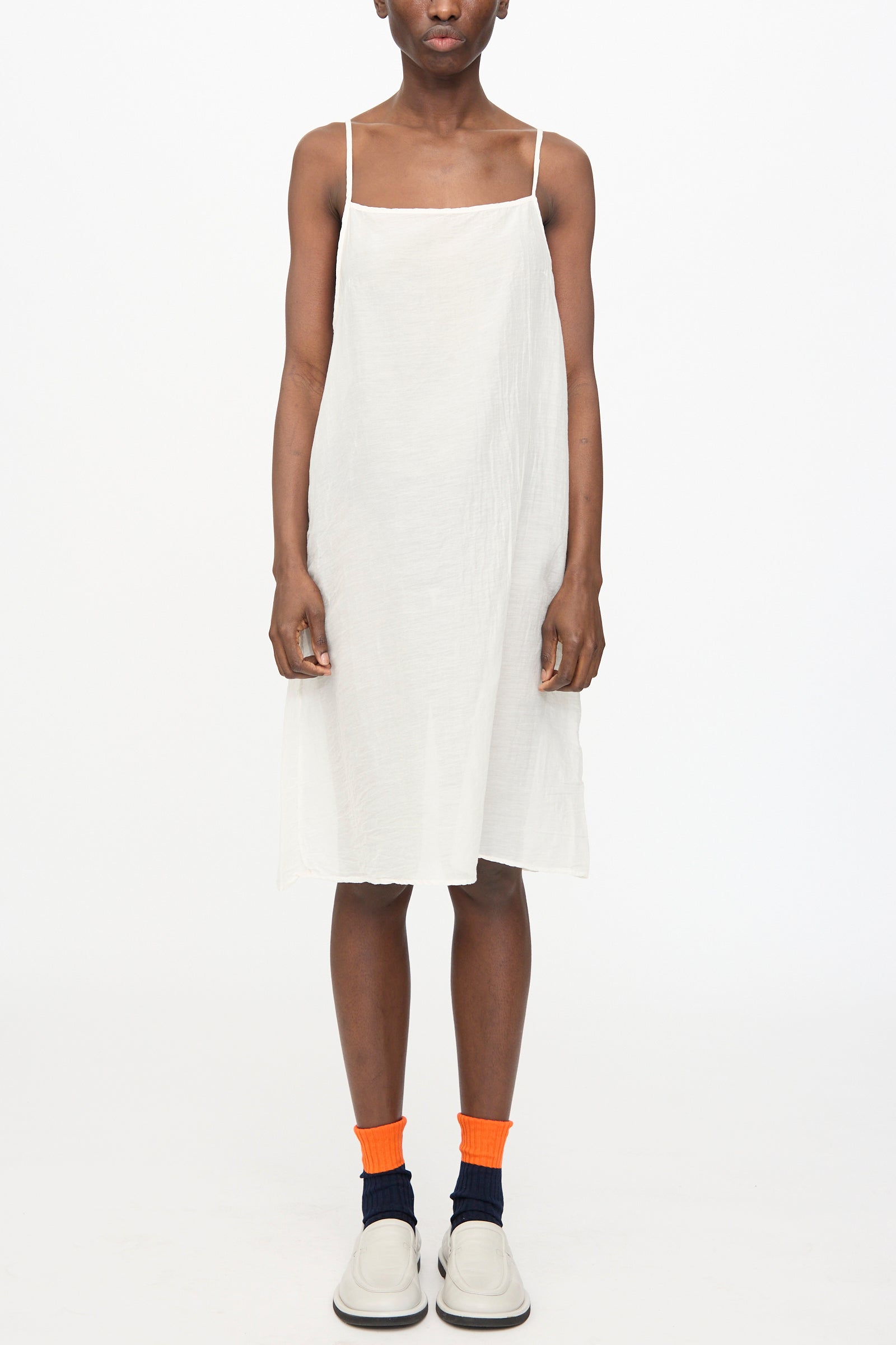 A person stands against a white background wearing the Black Crane Cami Dress in White, paired with orange and navy socks and white shoes.