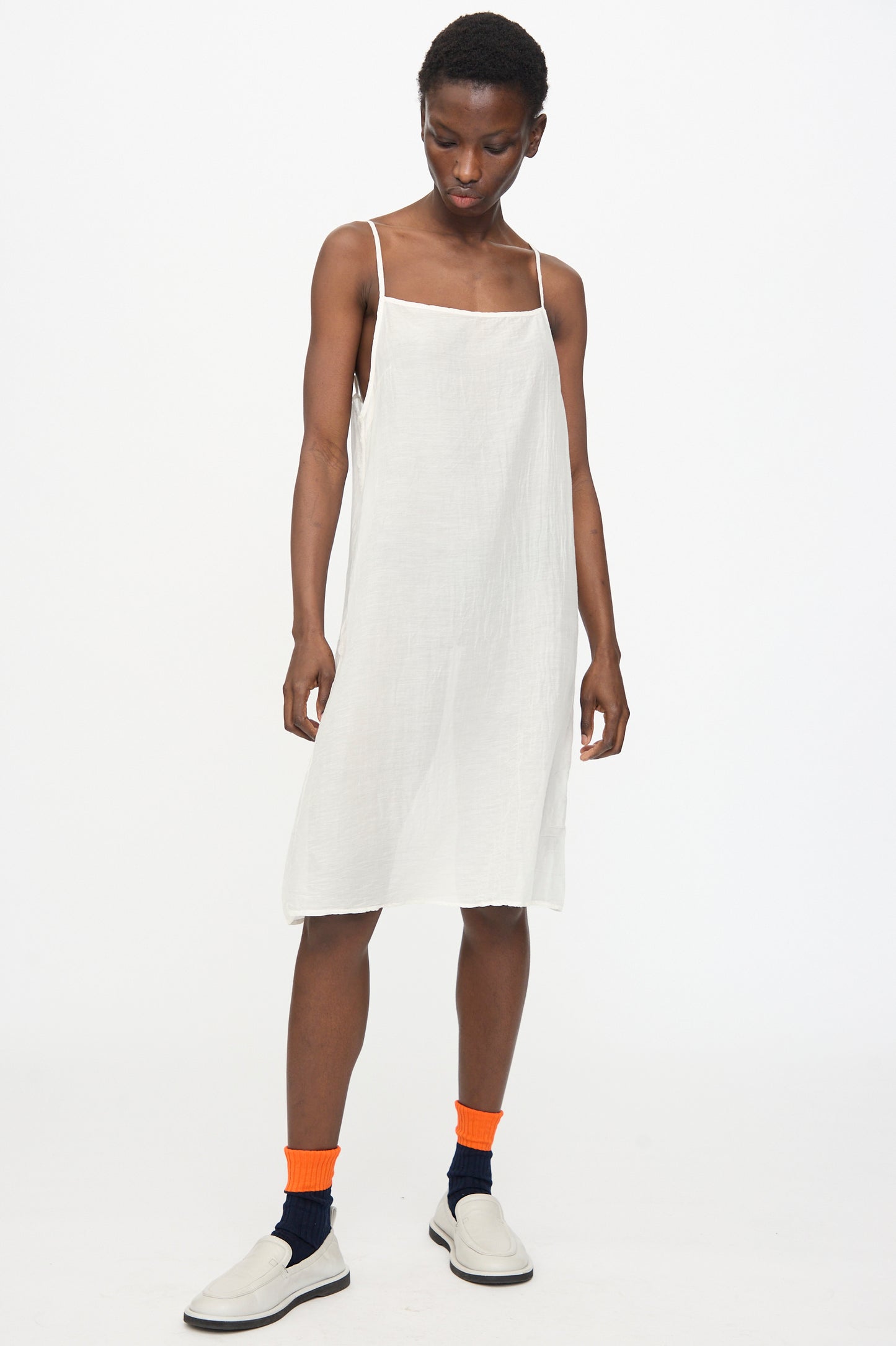A model wears the Black Crane Cami Dress in White with white slip-on shoes and layered orange over navy socks, standing against a plain white background.