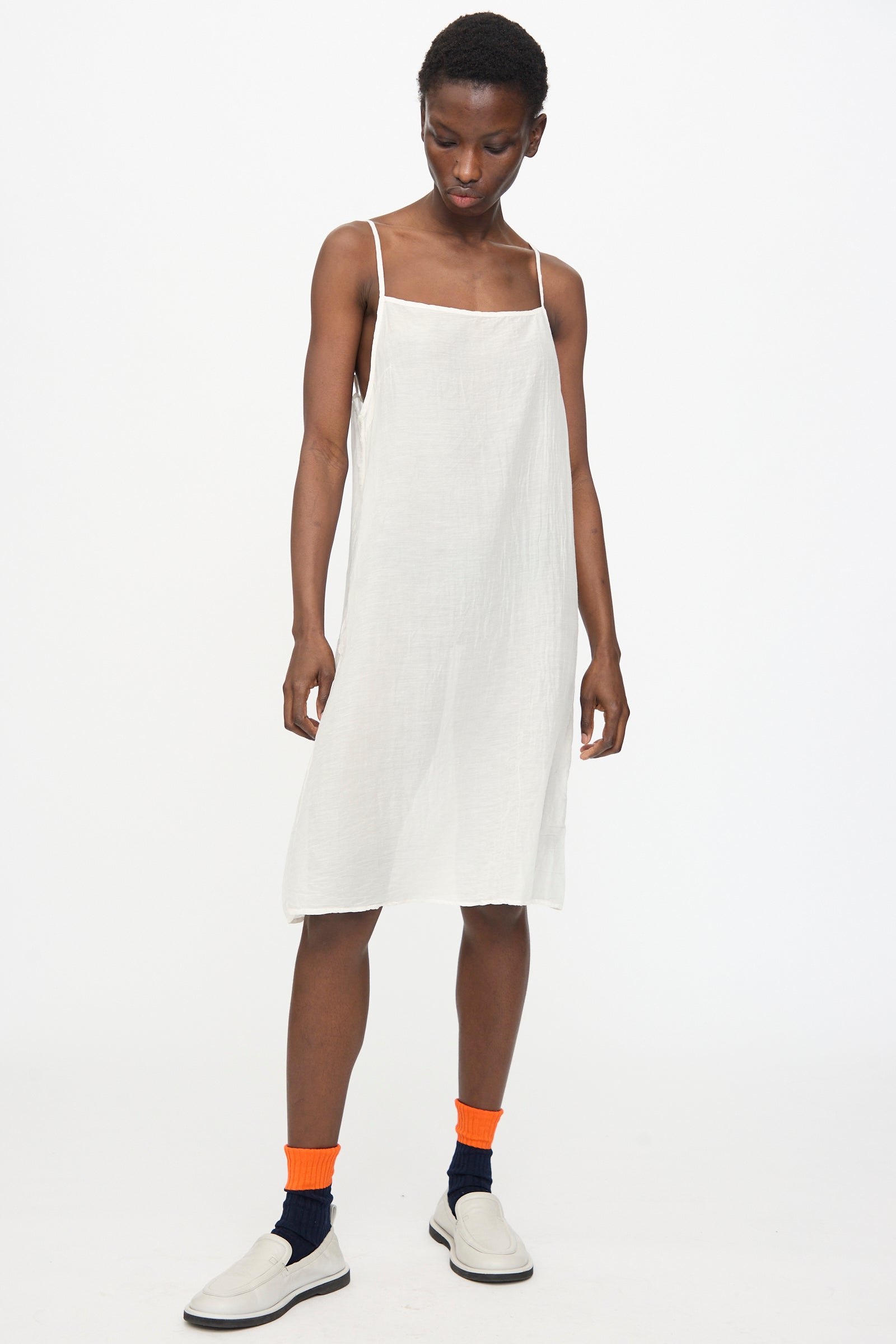 A model wears the Black Crane Cami Dress in White with white slip-on shoes and layered orange over navy socks, standing against a plain white background.