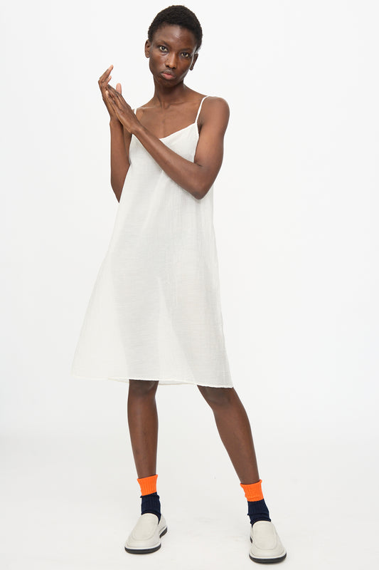 A person stands against a white background wearing the Black Crane Cami Dress in White, paired with orange socks and white slip-on shoes.