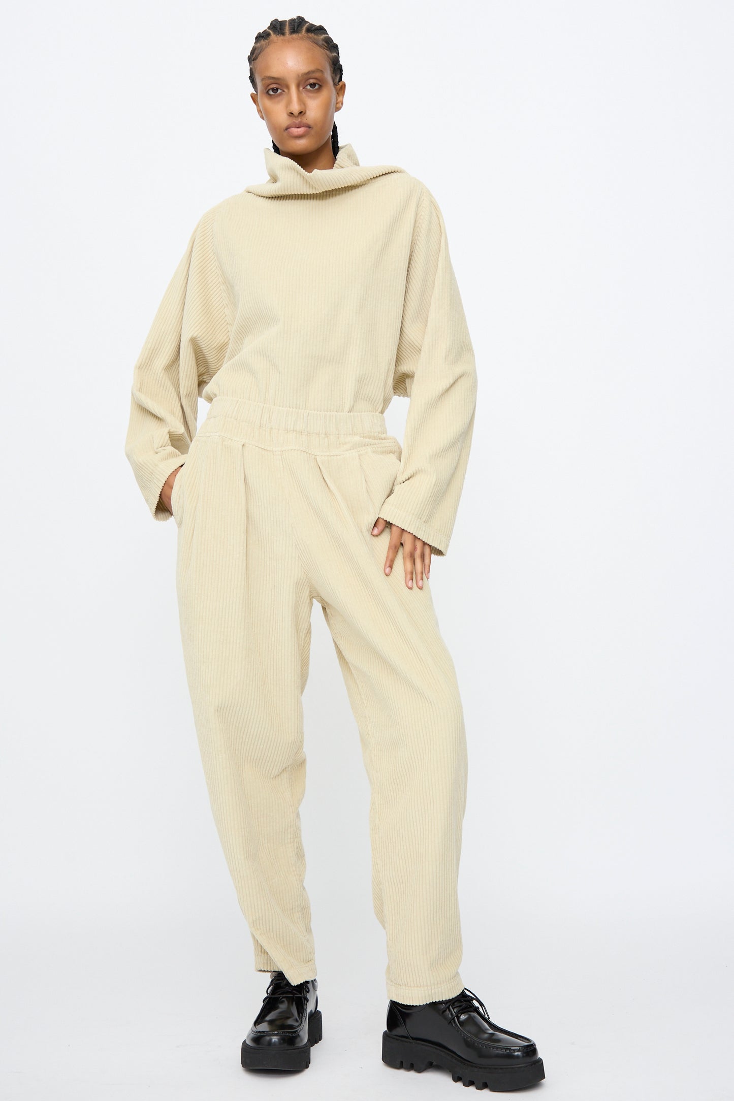 A person stands against a white background wearing Black Crane's Carpenter Pants in Ivory—loose, high-waisted cotton corduroy—and a relaxed fit, long-sleeve top, paired with black platform shoes.