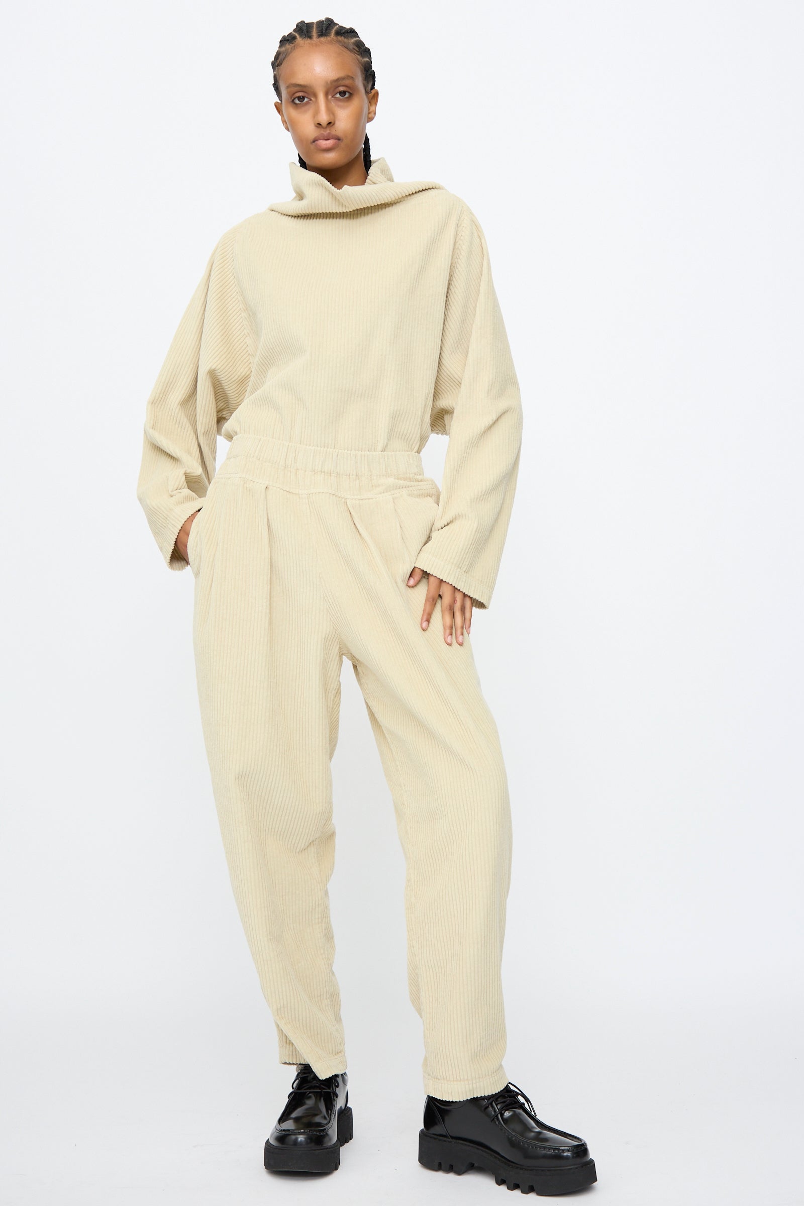 A person stands against a white background wearing Black Crane's Carpenter Pants in Ivory—loose, high-waisted cotton corduroy—and a relaxed fit, long-sleeve top, paired with black platform shoes.