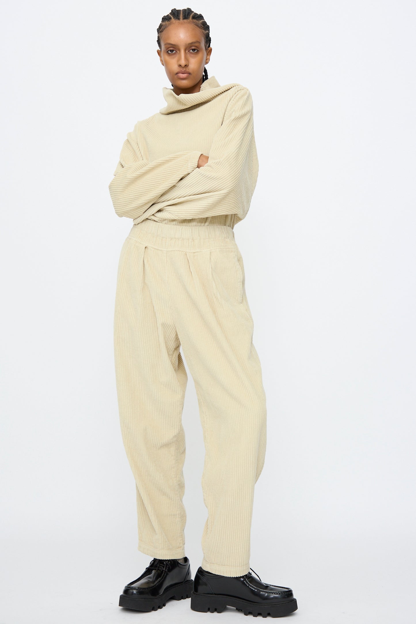 Person standing with arms crossed, wearing a loose beige turtleneck and Black Crane Carpenter Pants in Ivory with an elastic waist, paired with black platform shoes against a plain white background.