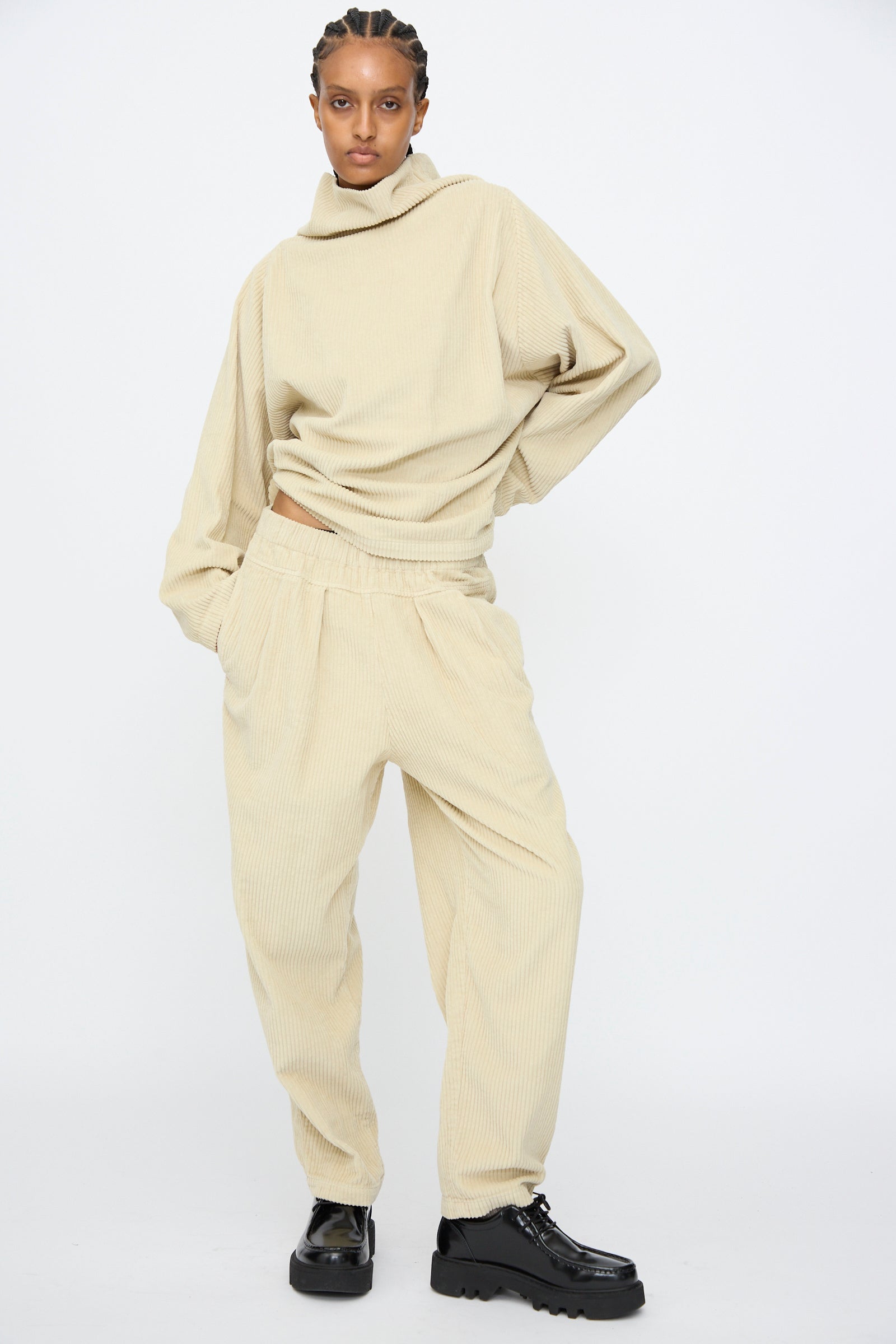 A person stands against a plain background wearing a loose, beige ribbed sweater and Black Crane Carpenter Pants in Ivory with an elastic waist, paired with black platform shoes.