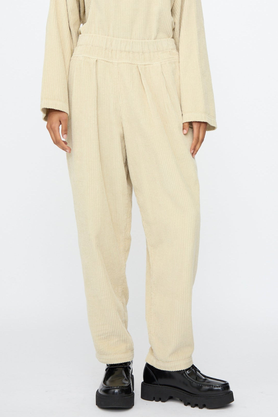 A person wears Black Crane Carpenter Pants in Ivory—relaxed fit, cotton corduroy with an elastic waist—paired with a matching long-sleeve top and black platform shoes against a plain background.