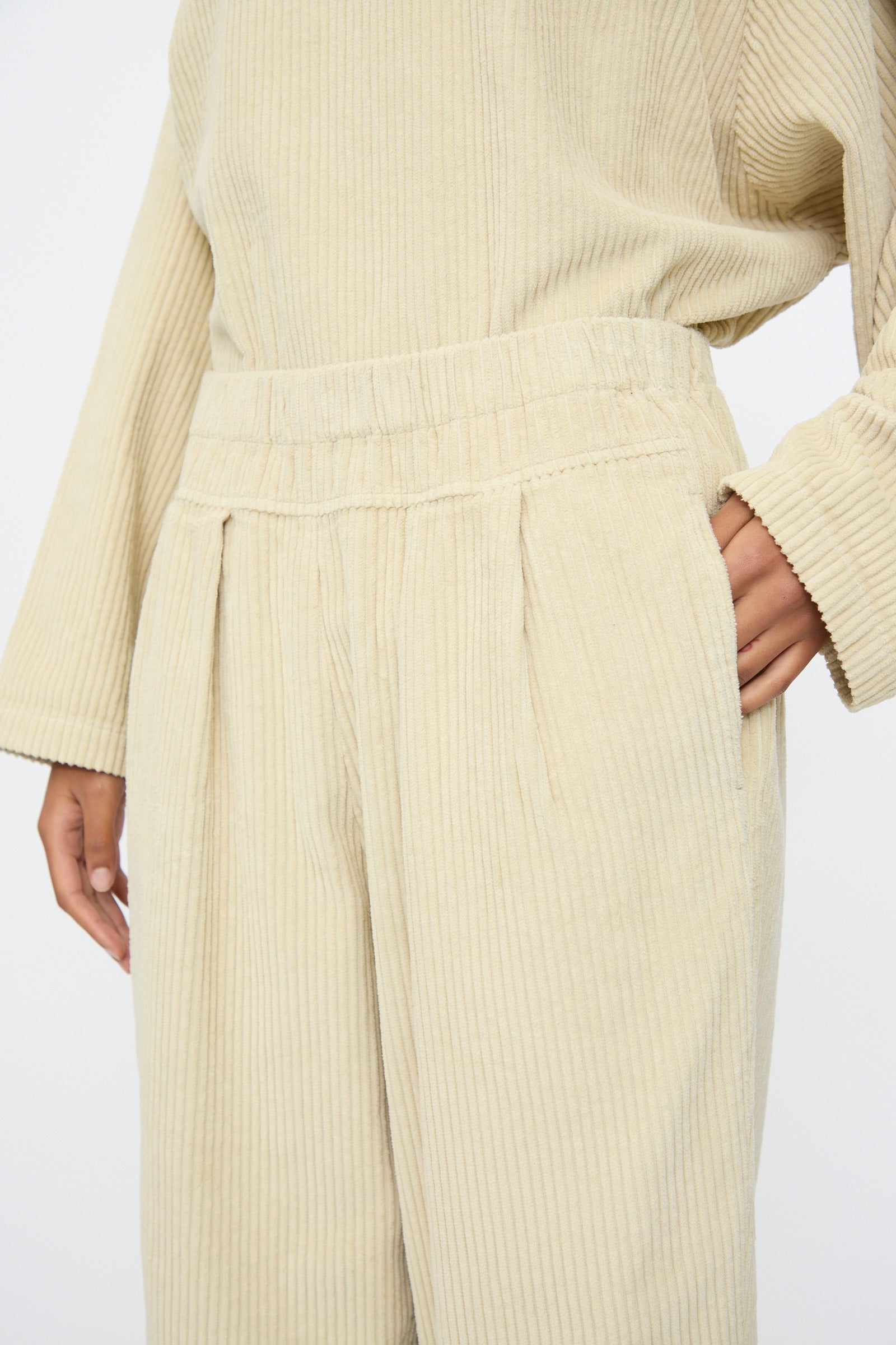 A person wears Black Crane’s Carpenter Pants in Ivory, styled with long sleeves; the relaxed-fit, high-waisted cotton corduroy design features one hand in the pocket.