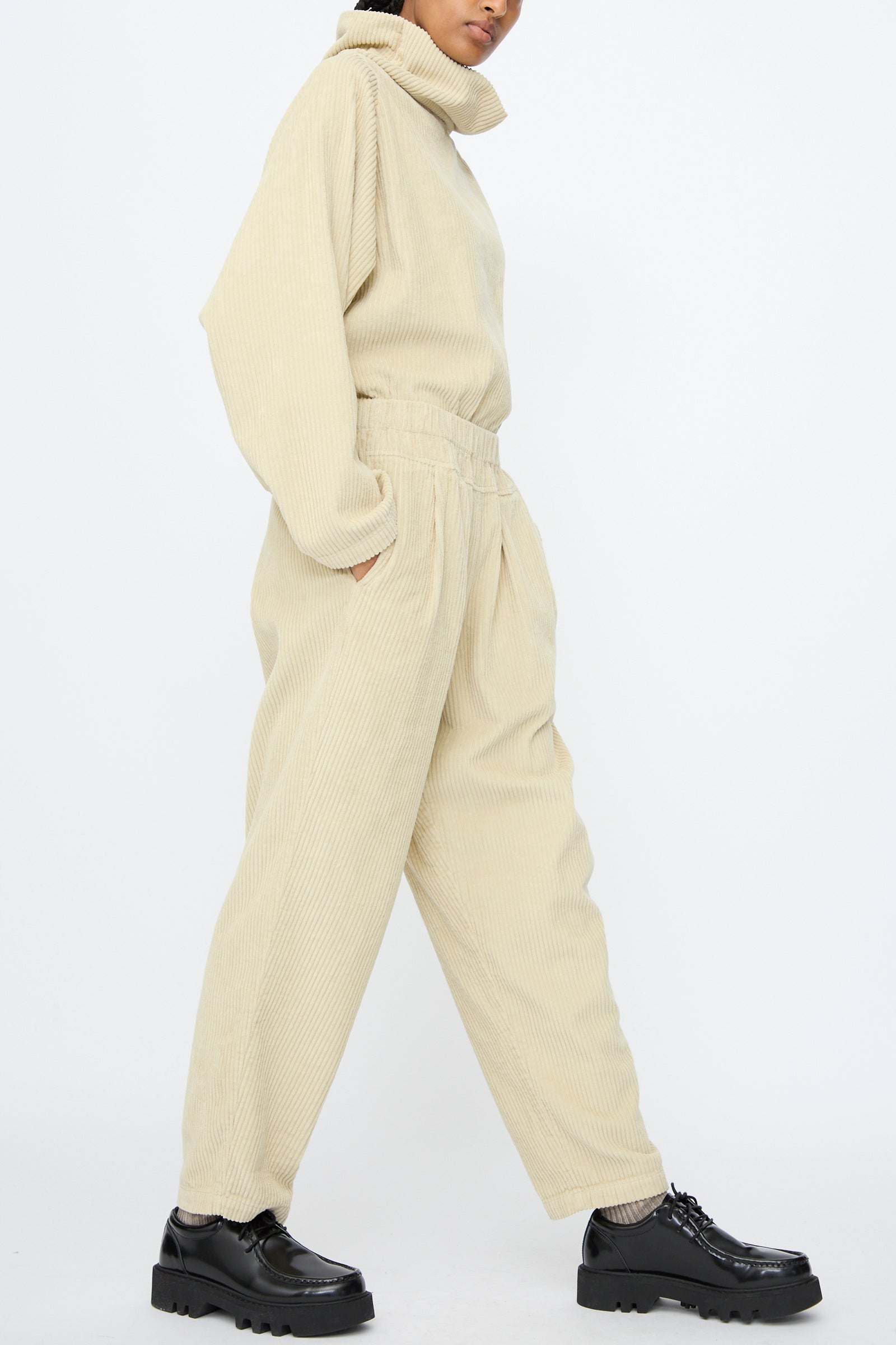 A person wearing Black Crane Carpenter Pants in Ivory, a beige relaxed fit corduroy set with long sleeves and elastic waist pants, paired with black platform shoes, stands against a plain white background.
